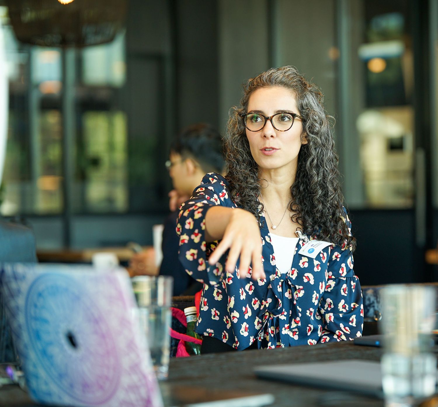 Woman with curly hair and glasses gesturing during a cafe meeting with laptop nearby.