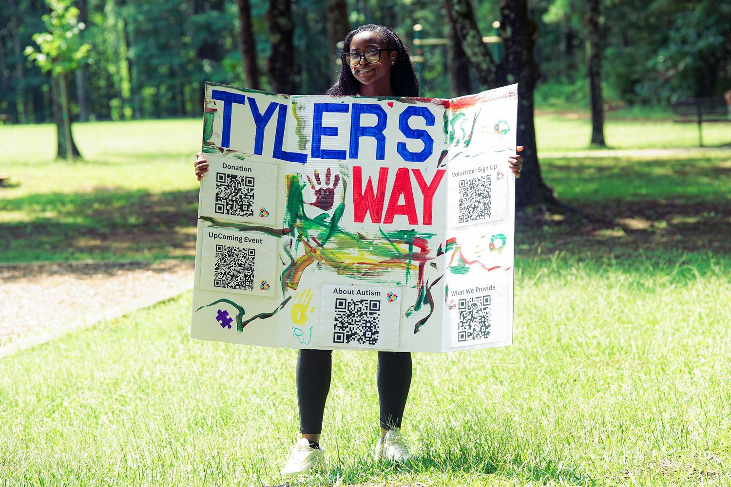 Young woman holds colorful Tyler's Way Foundation sign with QR codes in grassy park.