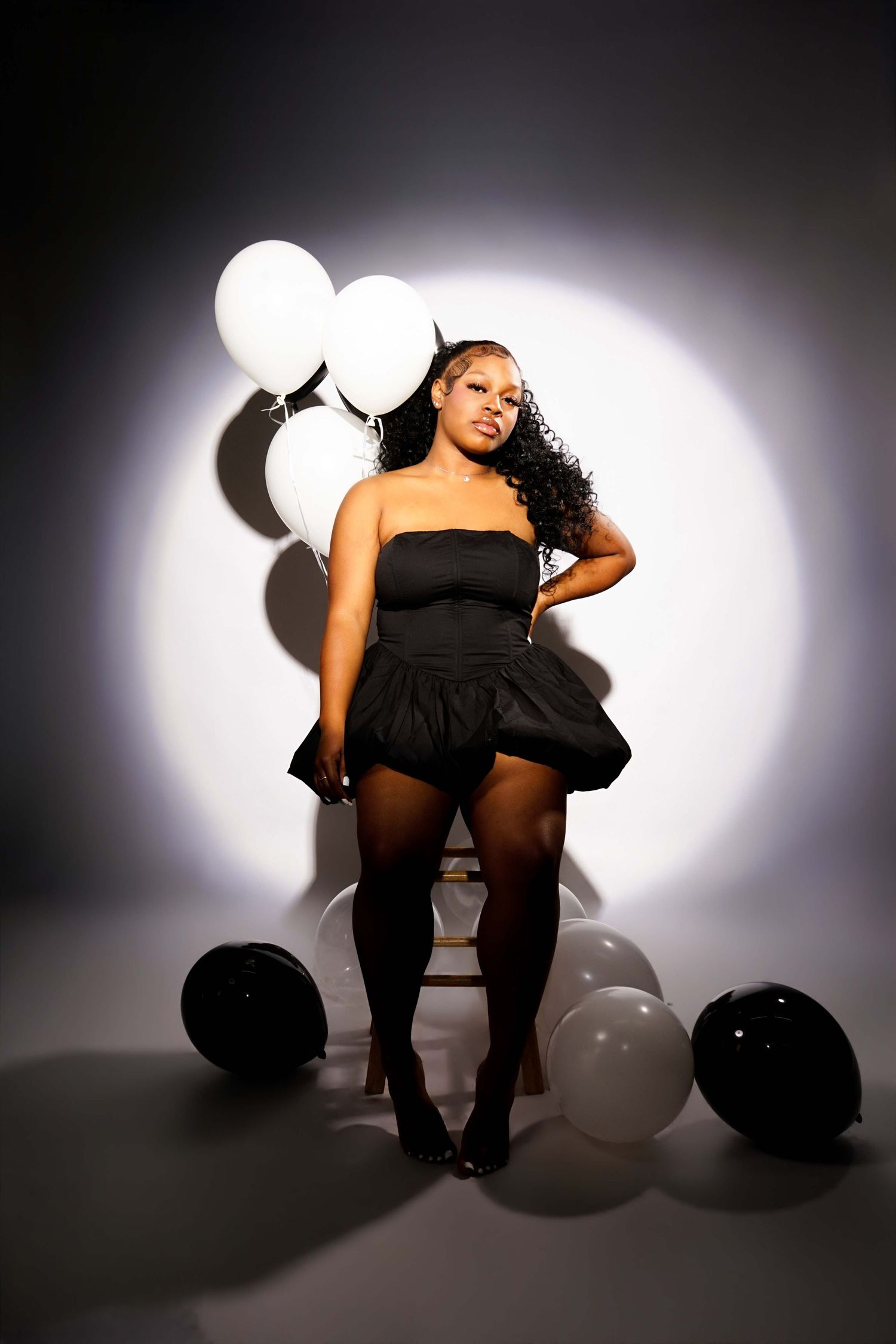 Woman in black strapless mini dress and tights posing with black and white balloons under dramatic studio spotlight.