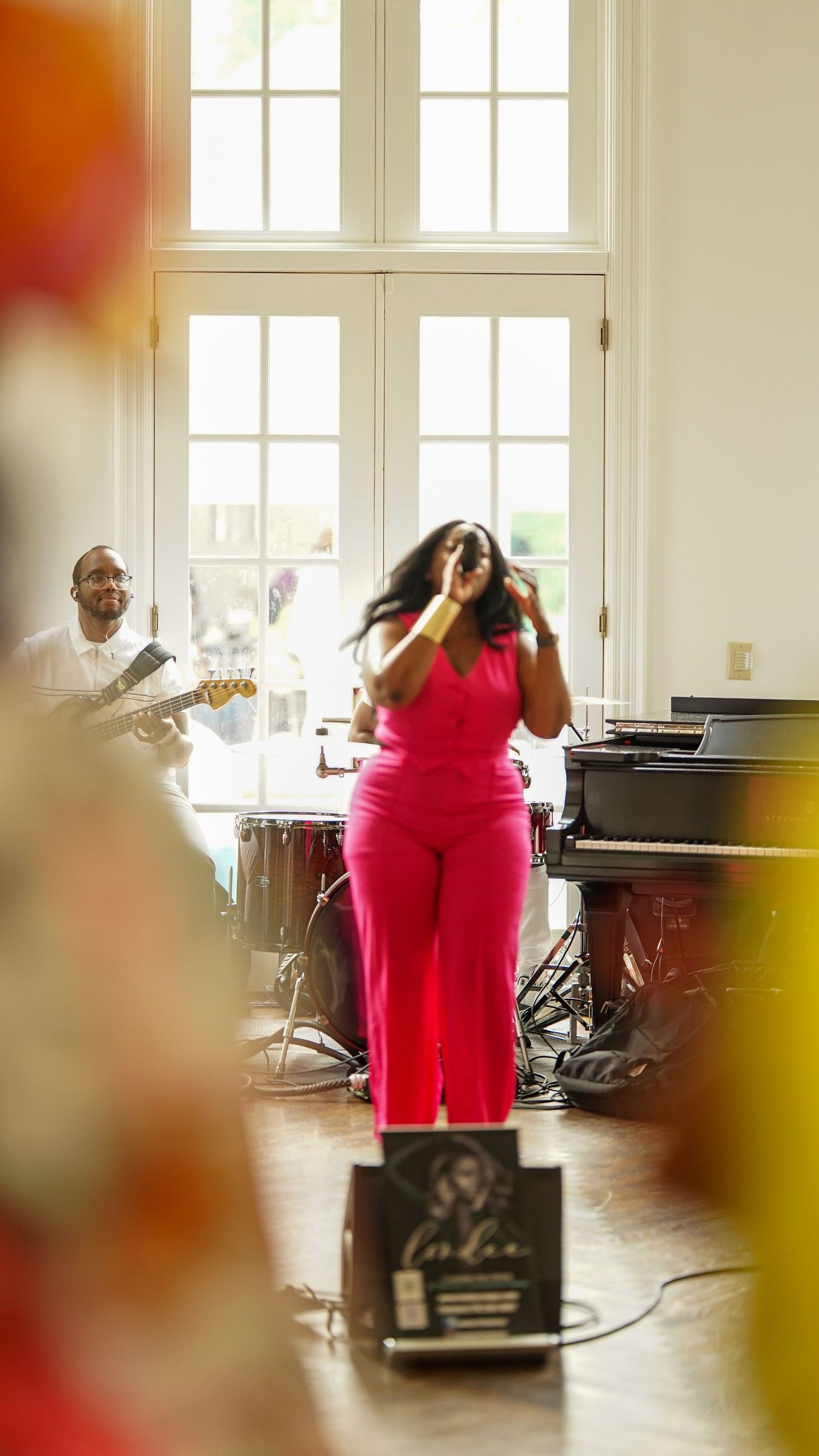Live musical performance in bright room with singer in red outfit and guitarist performing.