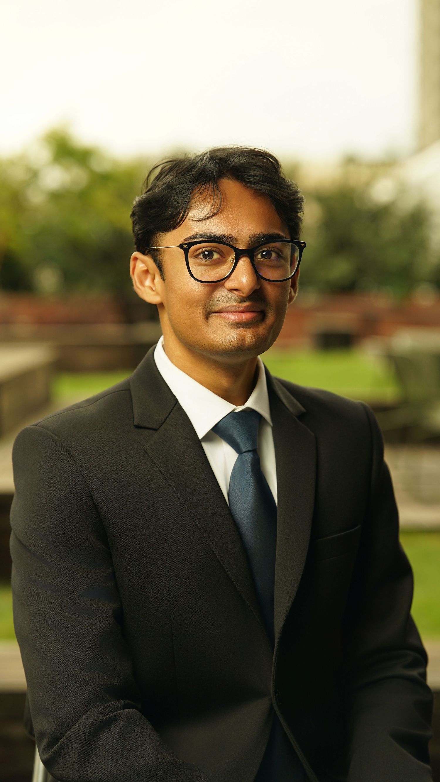 Professional headshot of person in black suit and blue tie with glasses against outdoor setting.