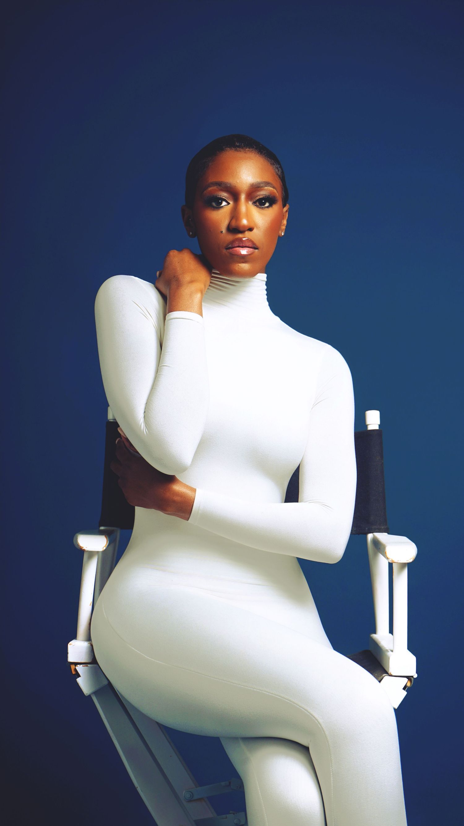 Model poses in fitted white turtleneck outfit against navy blue backdrop.