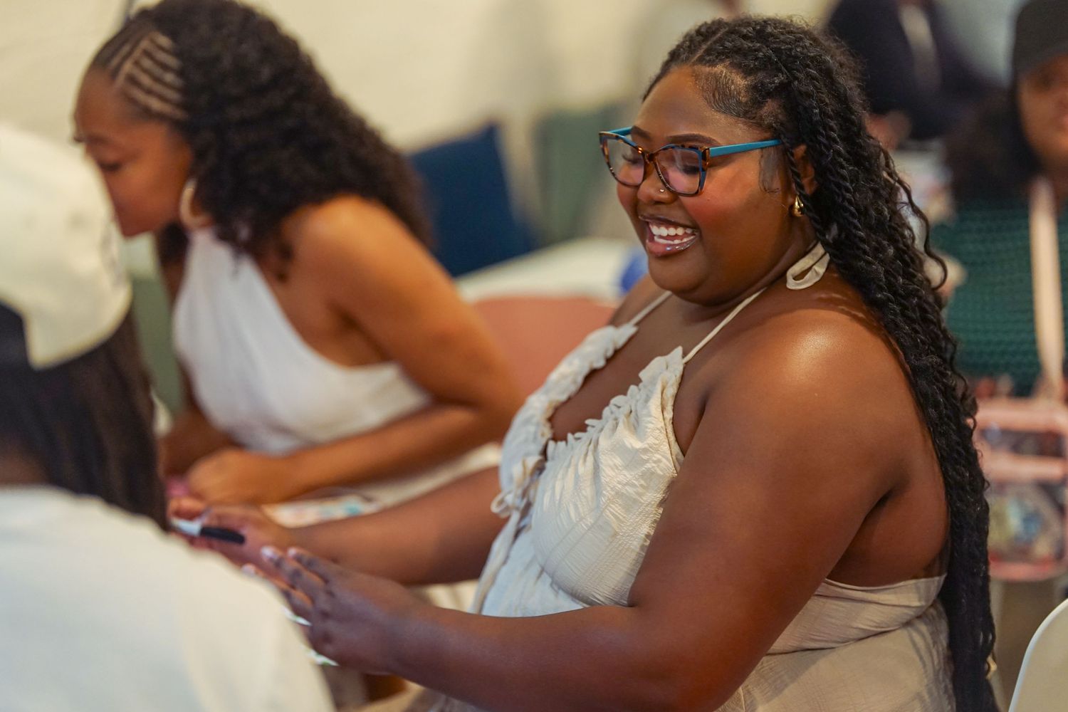 Young Black woman with glasses and braids smiling warmly during an indoor community event.