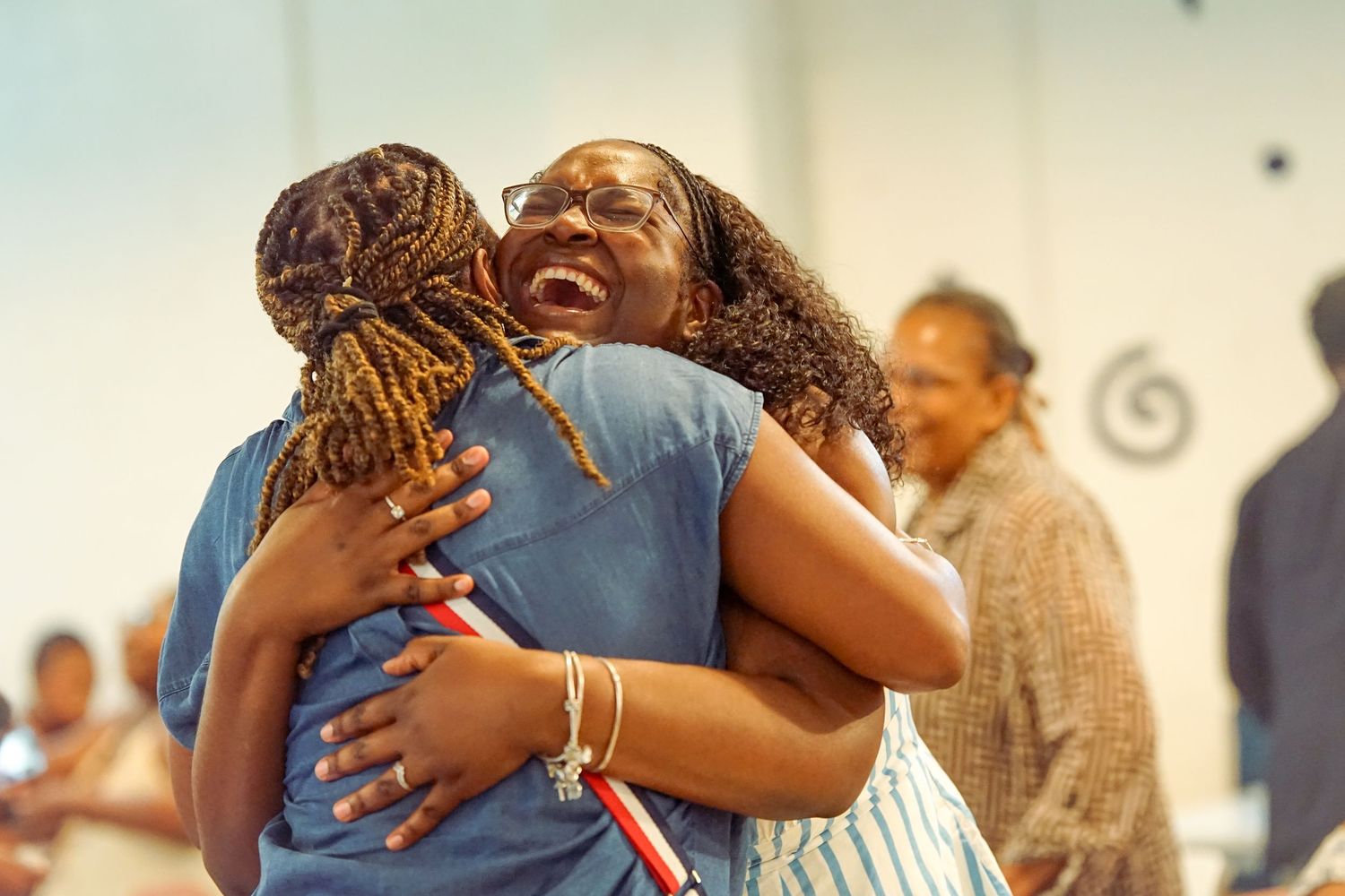 Two women share a joyful embrace, laughing and hugging warmly at an indoor gathering event.
