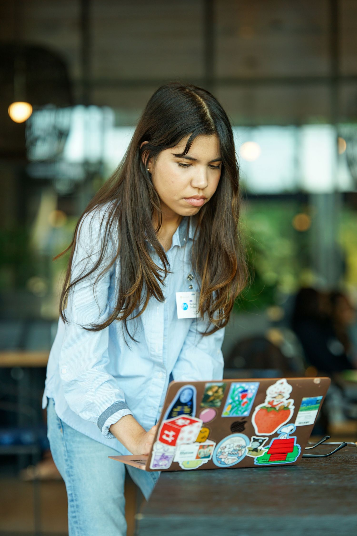 Young woman in blue shirt looking at sticker-covered laptop at a standing desk.