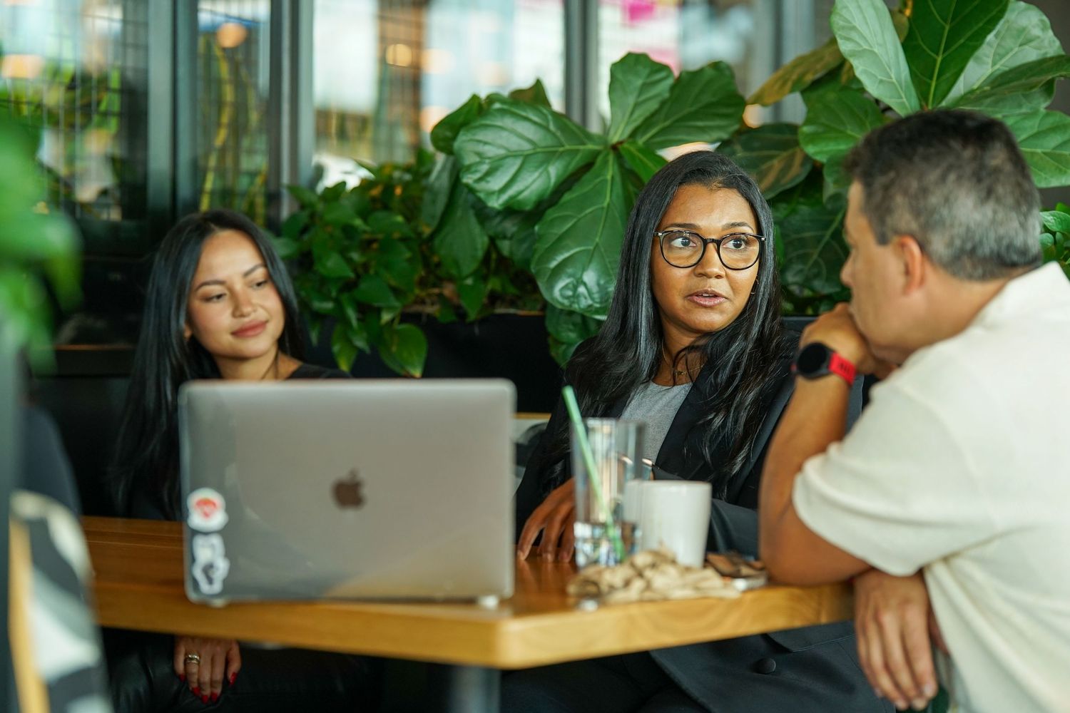 Three colleagues collaborate around a laptop at a cafe table surrounded by tropical plants.