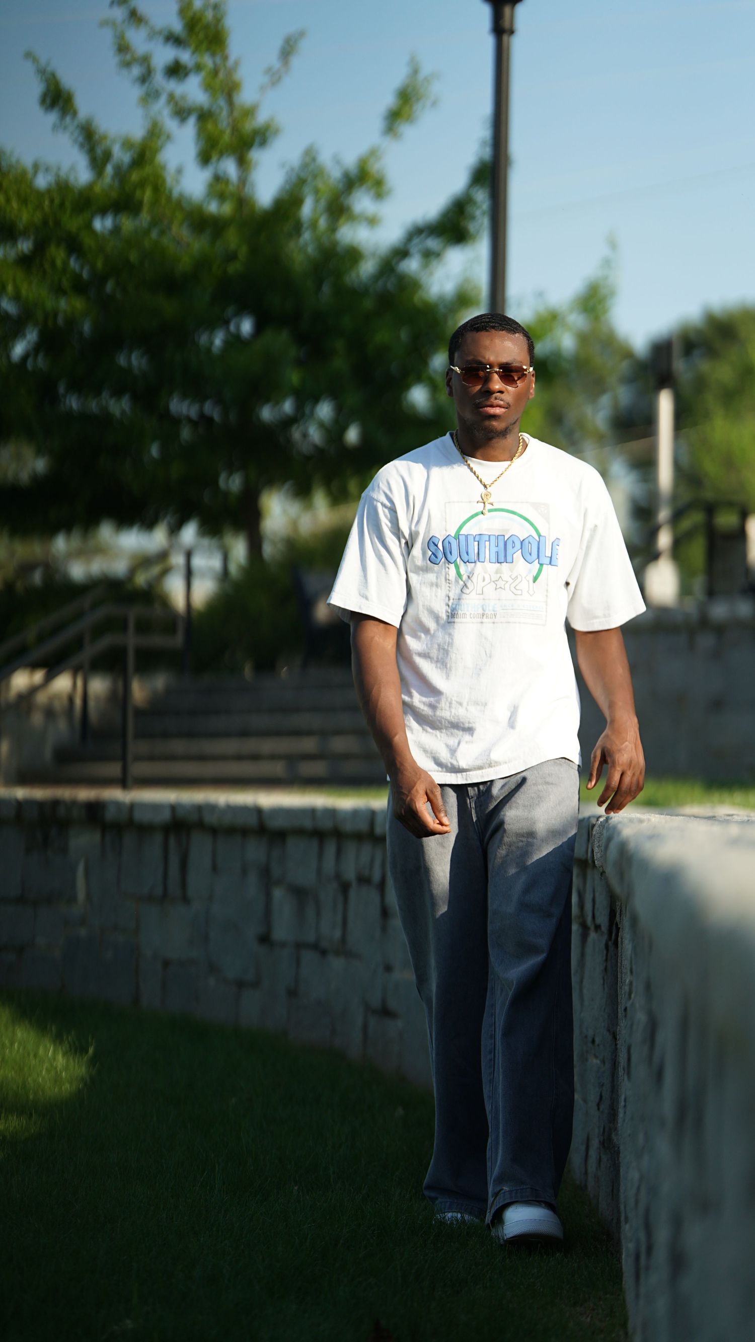 Casual portrait of person in white graphic tee and jeans standing against stone wall outdoors.