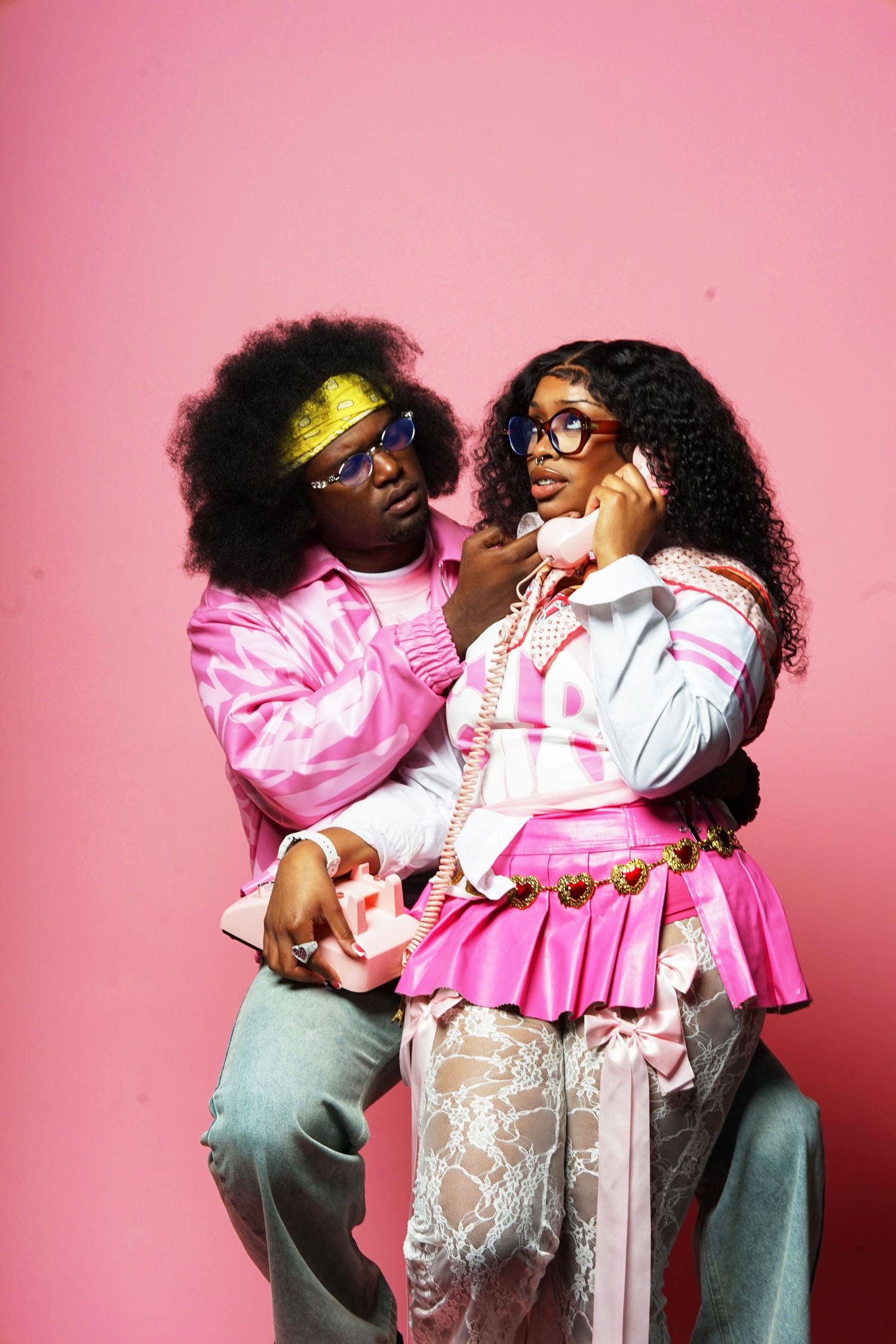 Two people in pink and white outfits pose playfully against pink backdrop.