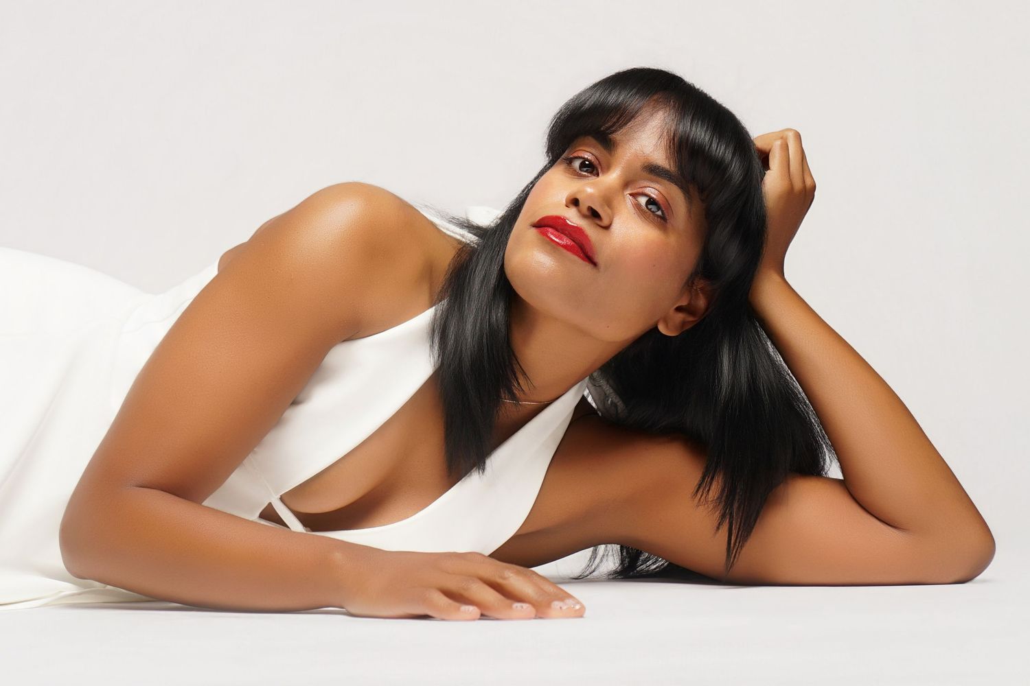 Young woman in white dress lying down, posing elegantly with red lips and dark hair against white background.