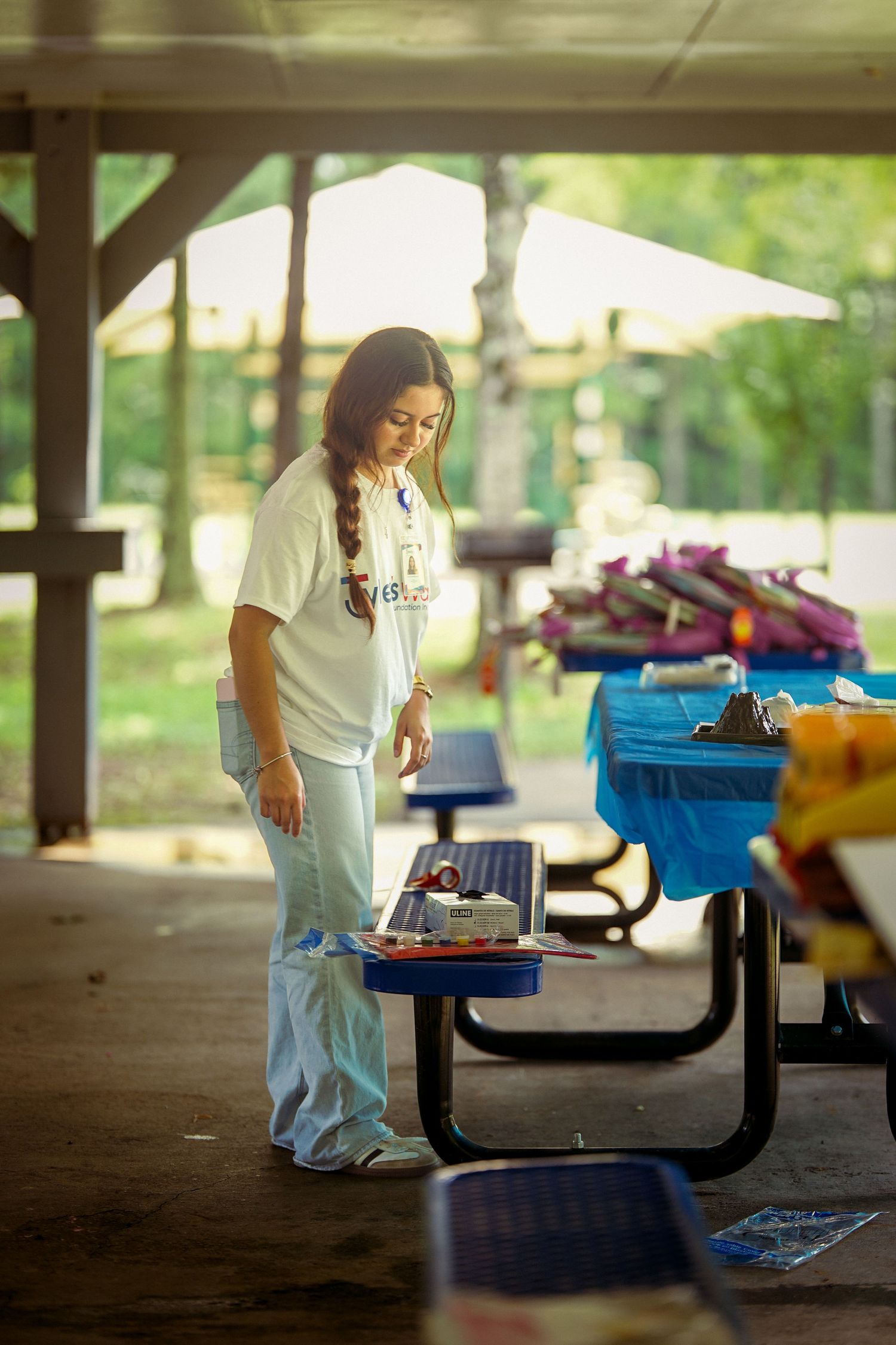 A young woman with a braid examines craft supplies on a picnic table under an outdoor pavilion.