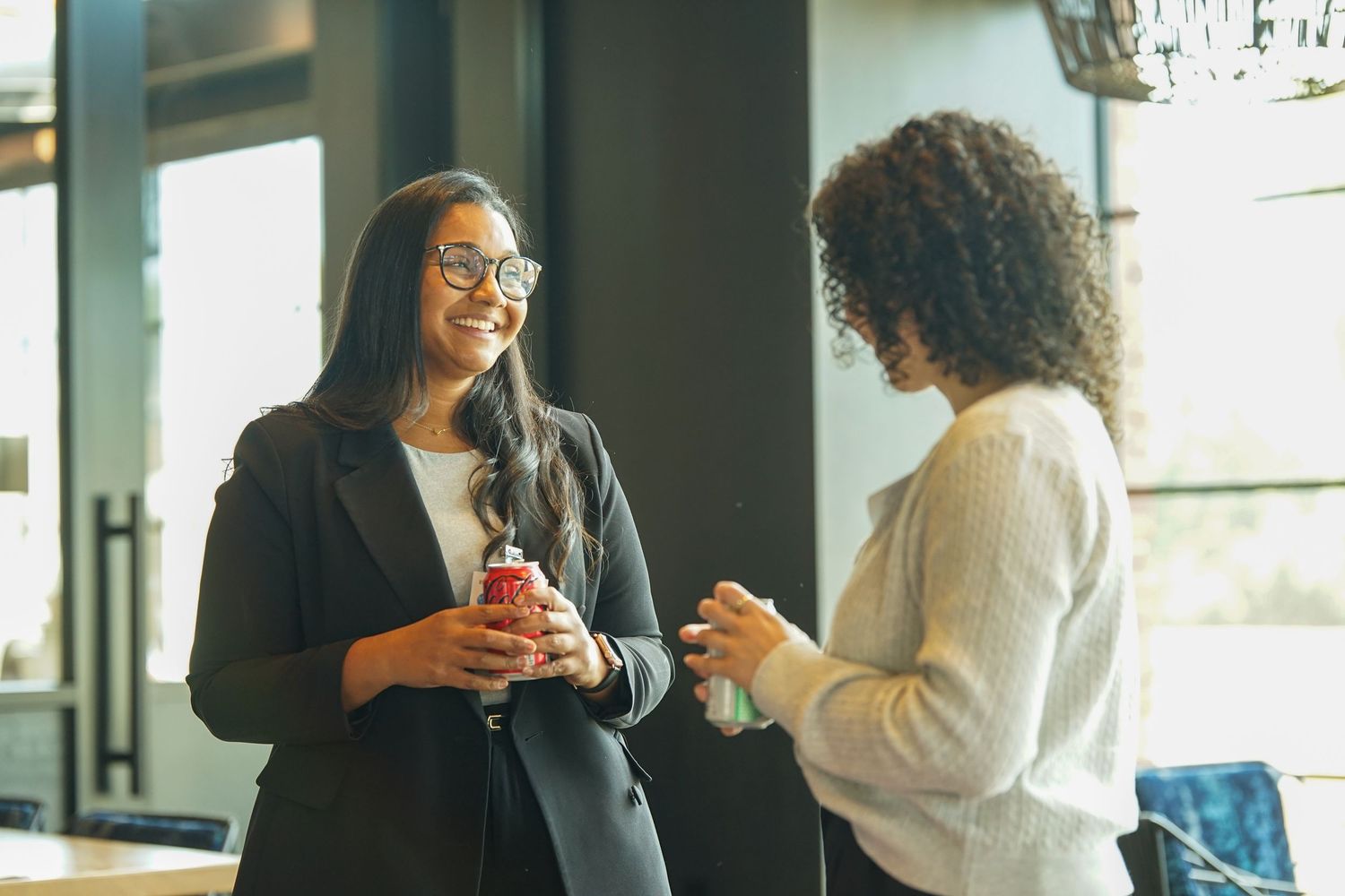 Two professional women smile and chat while holding drinks at a business networking event.