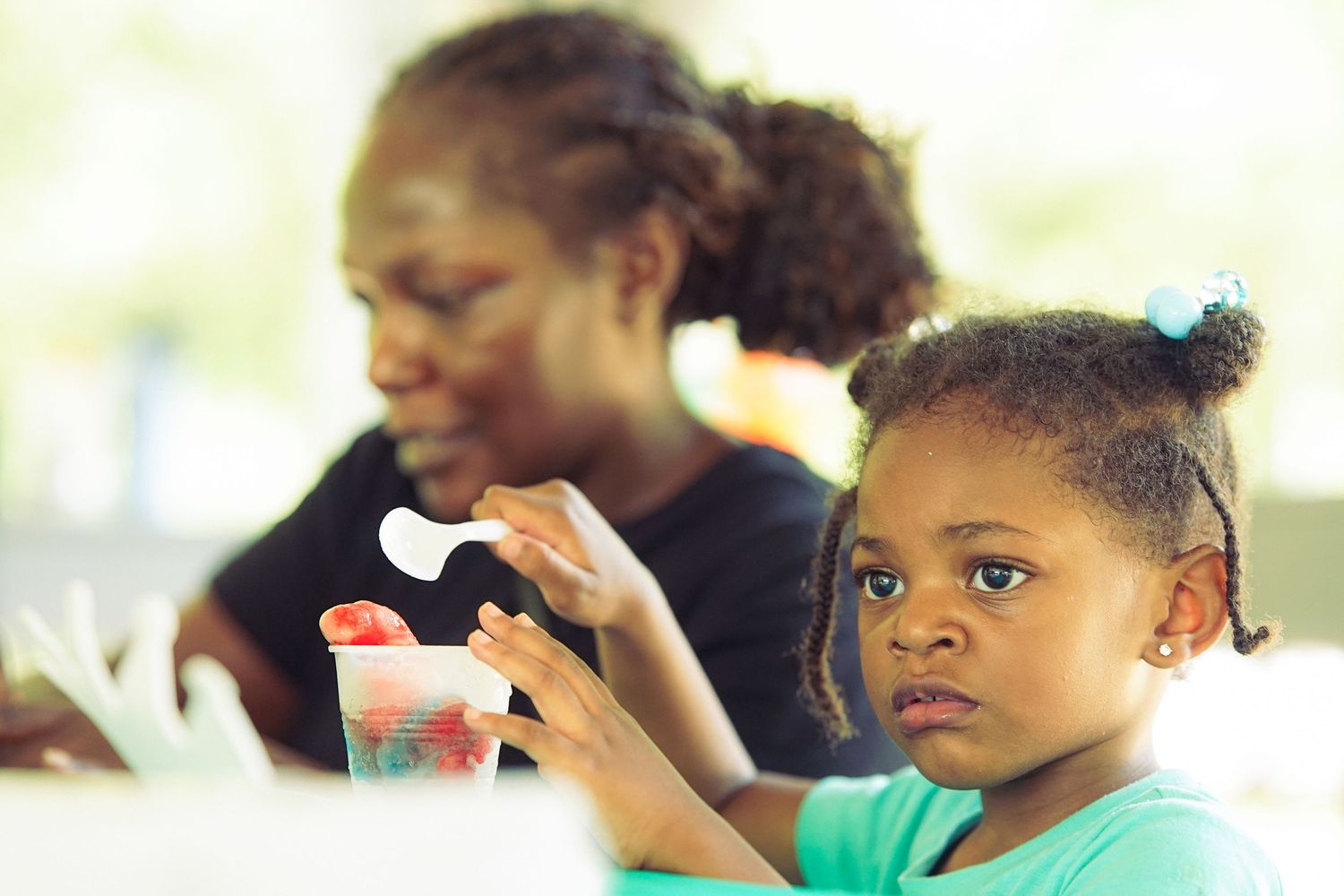 Young girl in teal shirt holds a drink with straw while woman sits behind her at an outdoor table.