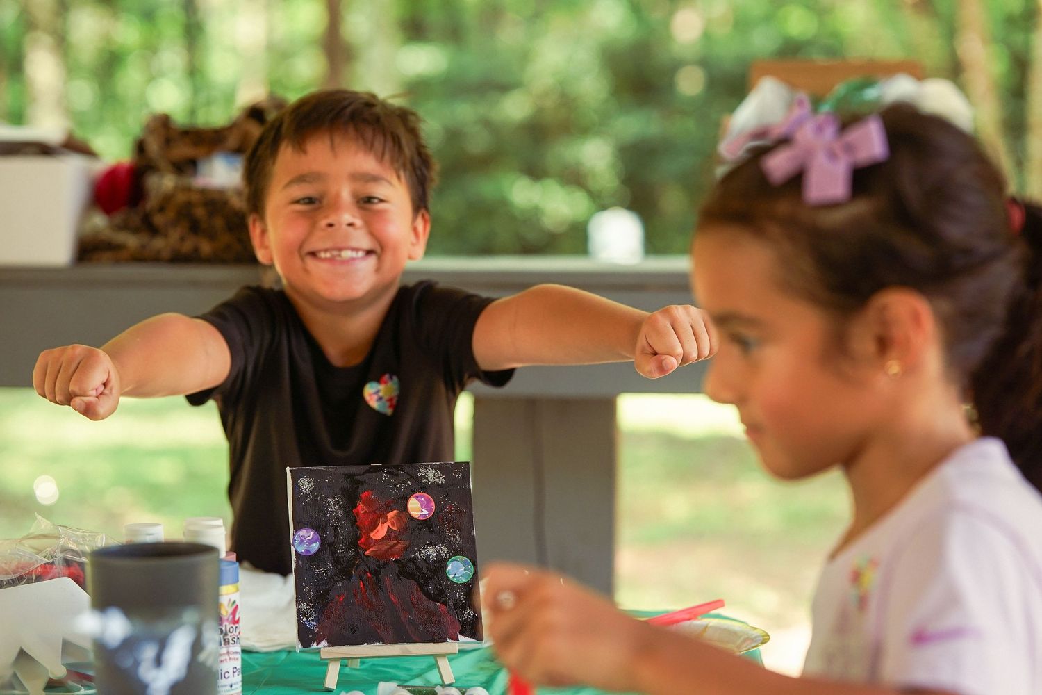 Two children smile and craft art projects at an outdoor picnic table surrounded by lush green trees.
