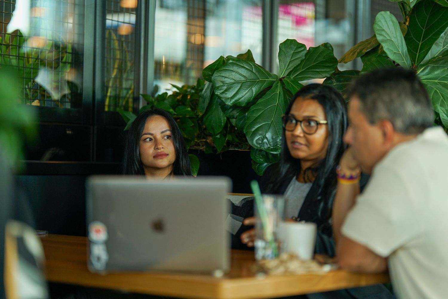 Two young women and a man collaborate over a laptop in a plant-filled cafe setting.