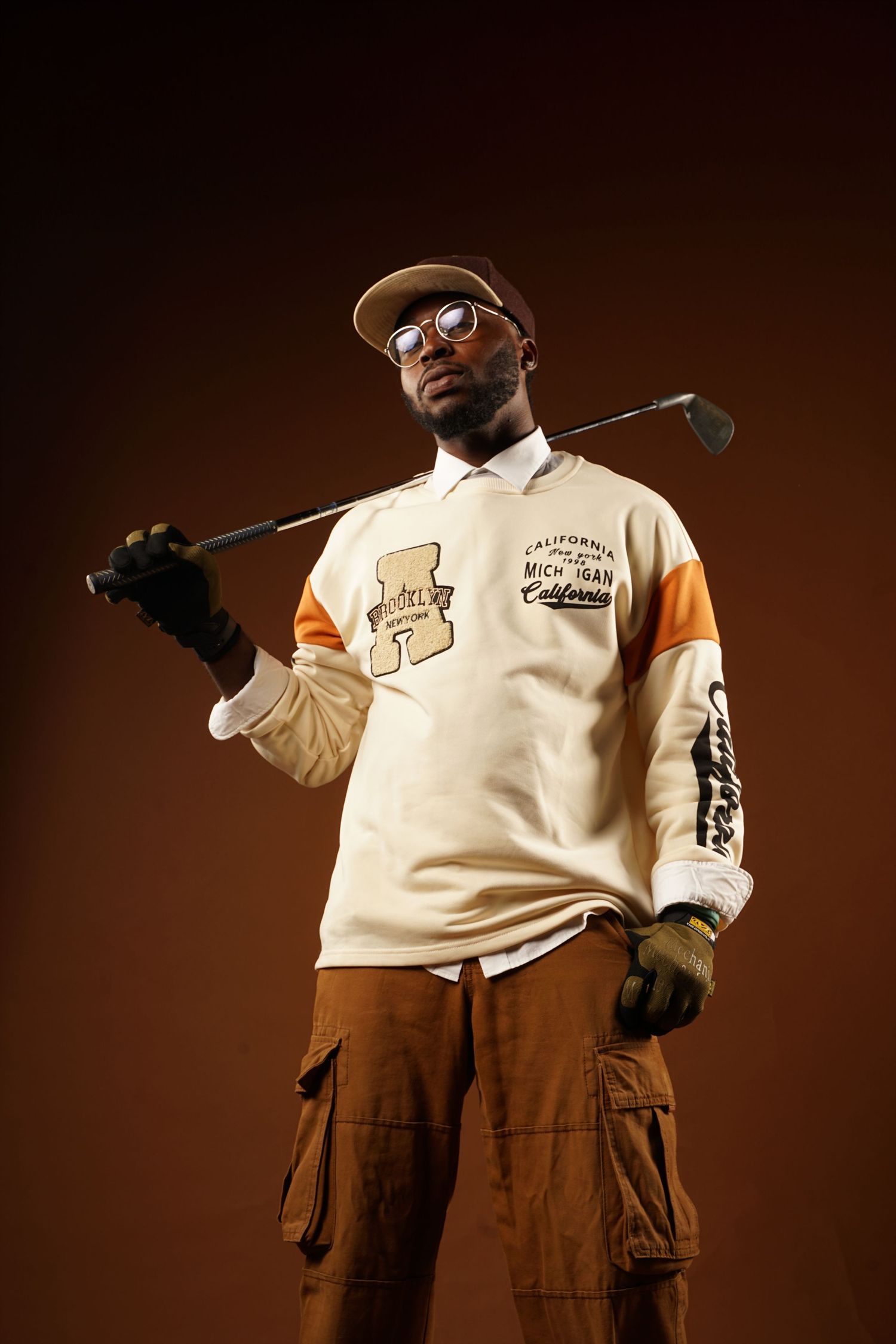 Person wearing a cream colored sweater and brown cargo pants poses against a dark brown backdrop.