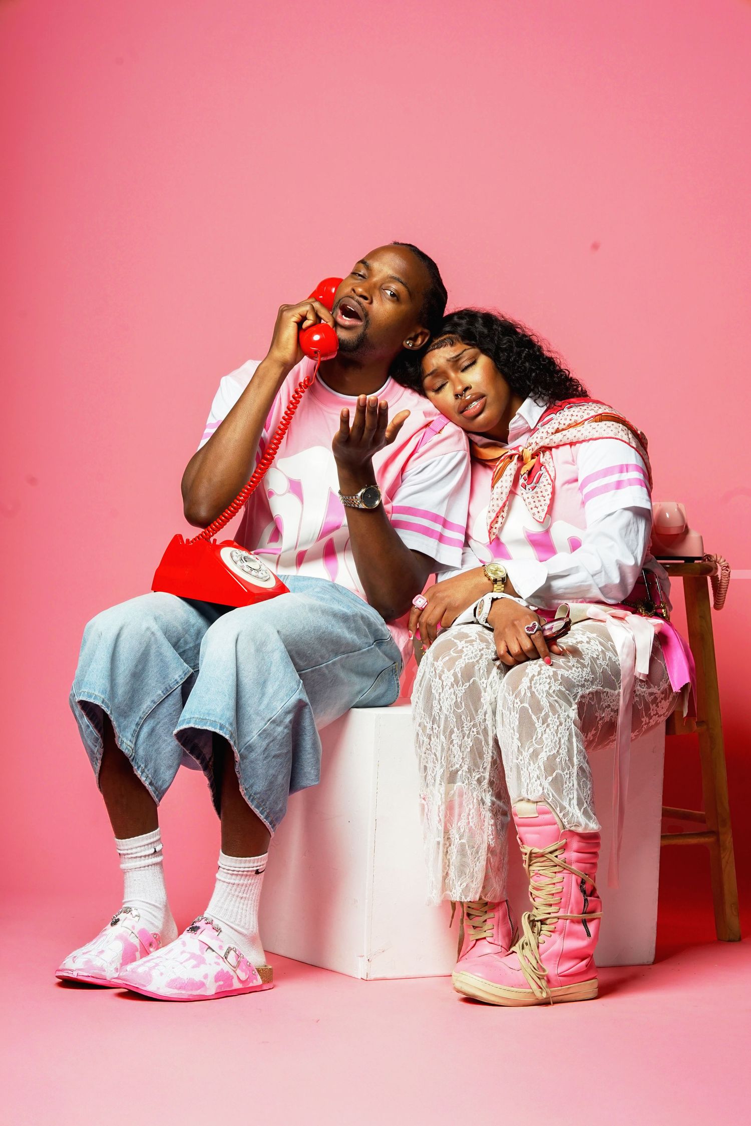 Models relaxing on white blocks wearing casual streetwear against pink backdrop.