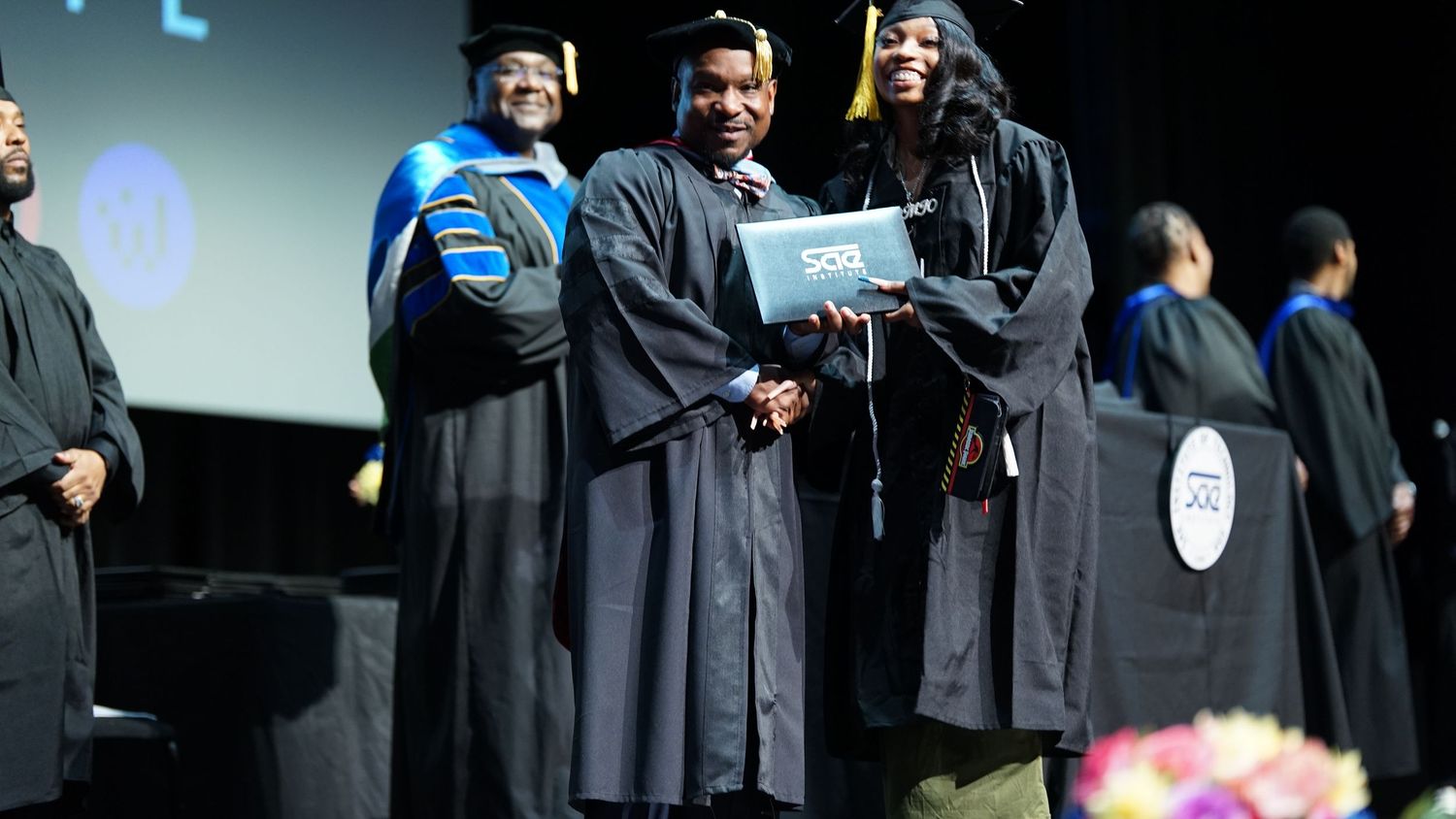 Wide shot of graduation ceremony with graduates in black robes on stage.