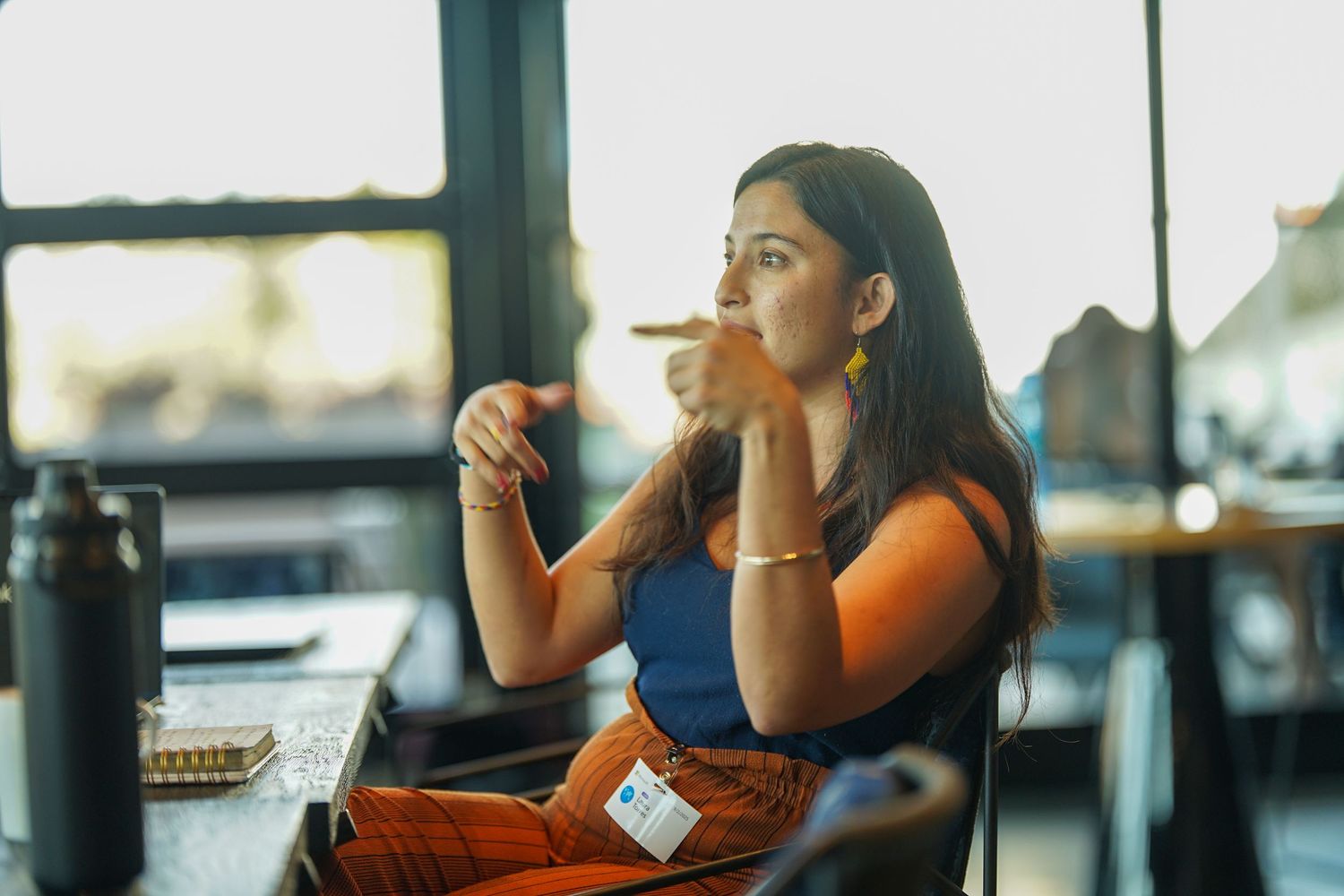 Young woman sitting in a cafe, thoughtfully sipping a drink, wearing a blue top and orange pants.