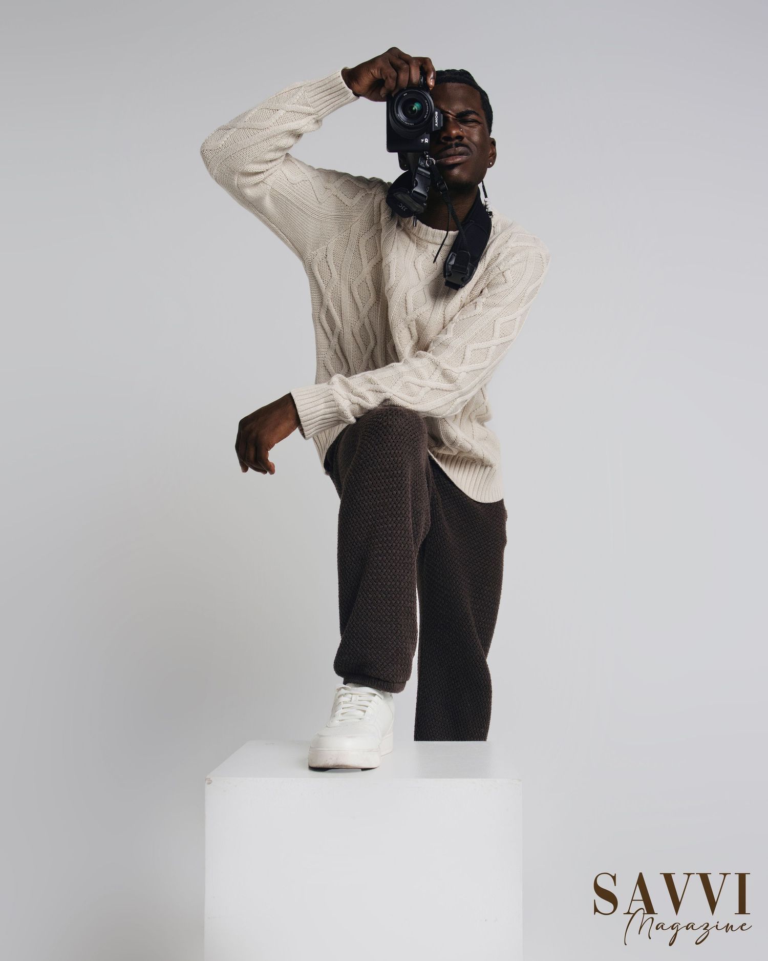 Fashion model in cream sweater and dark pants poses dynamically on white pedestal.