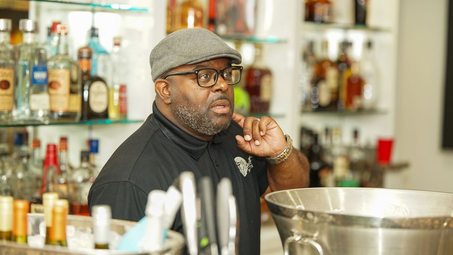Bartender in black attire and cap stands behind bar counter with liquor bottles in background.