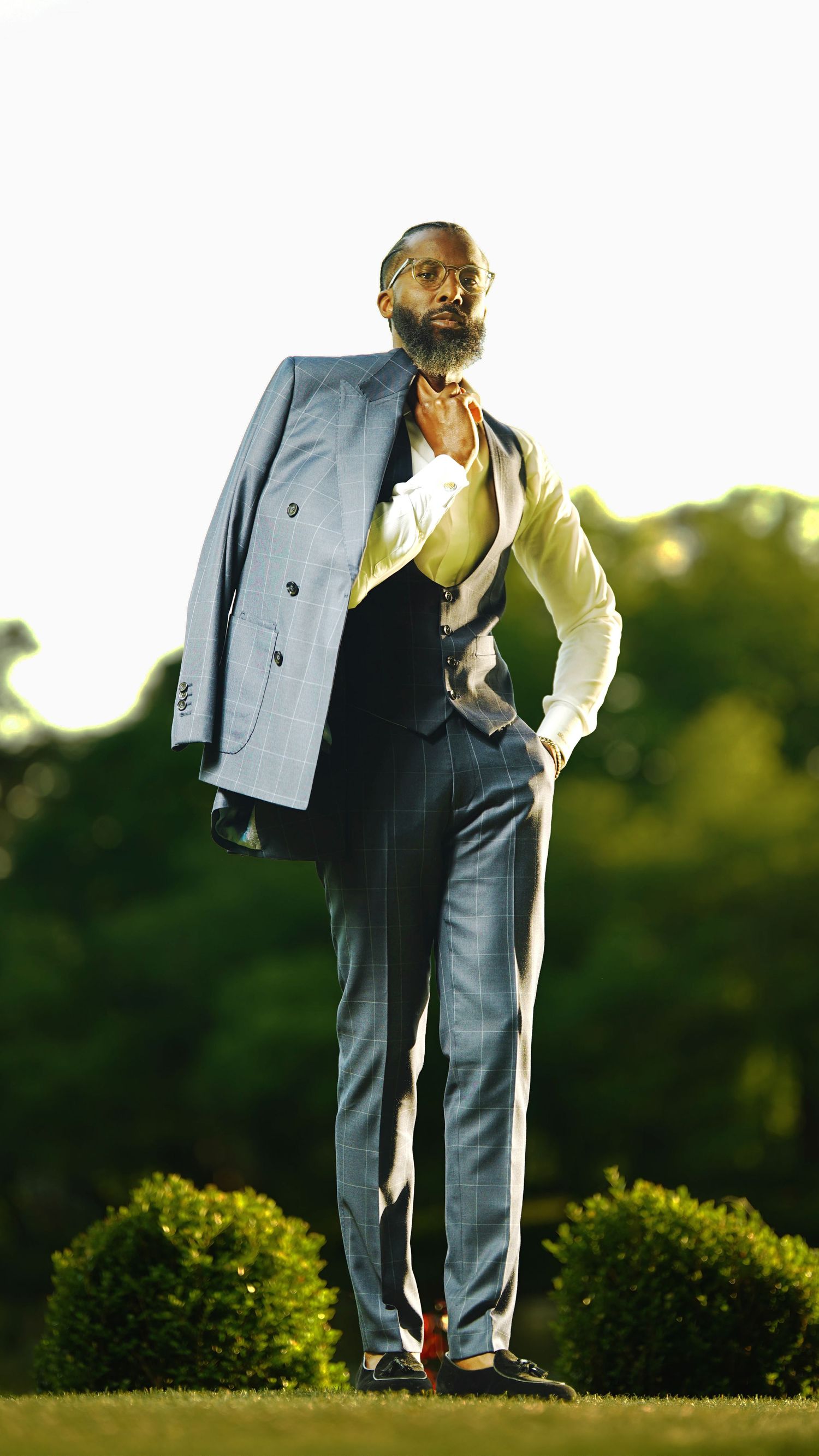 Person in a gray suit and yellow shirt poses playfully in an outdoor setting with greenery in the background.
