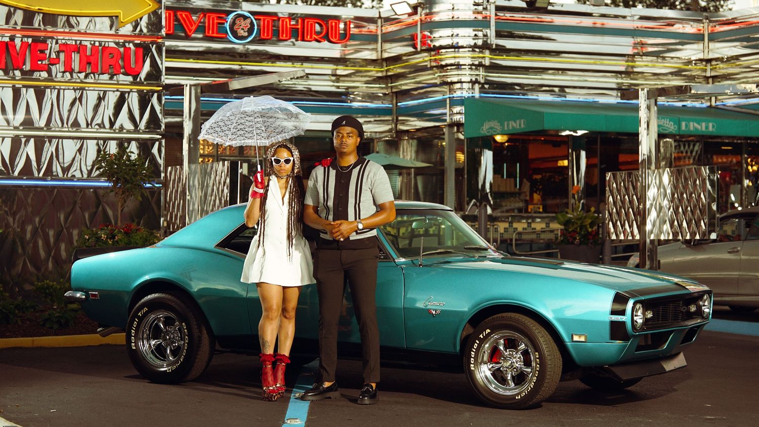 A stylish couple poses with a vintage teal Camaro at a retro drive-in setting.