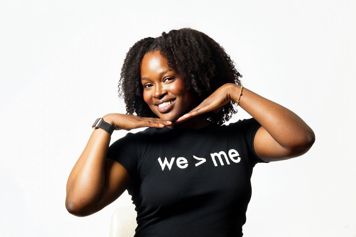 Woman smiling confidently wearing a black t-shirt with 'we > me' slogan, posing against white background.