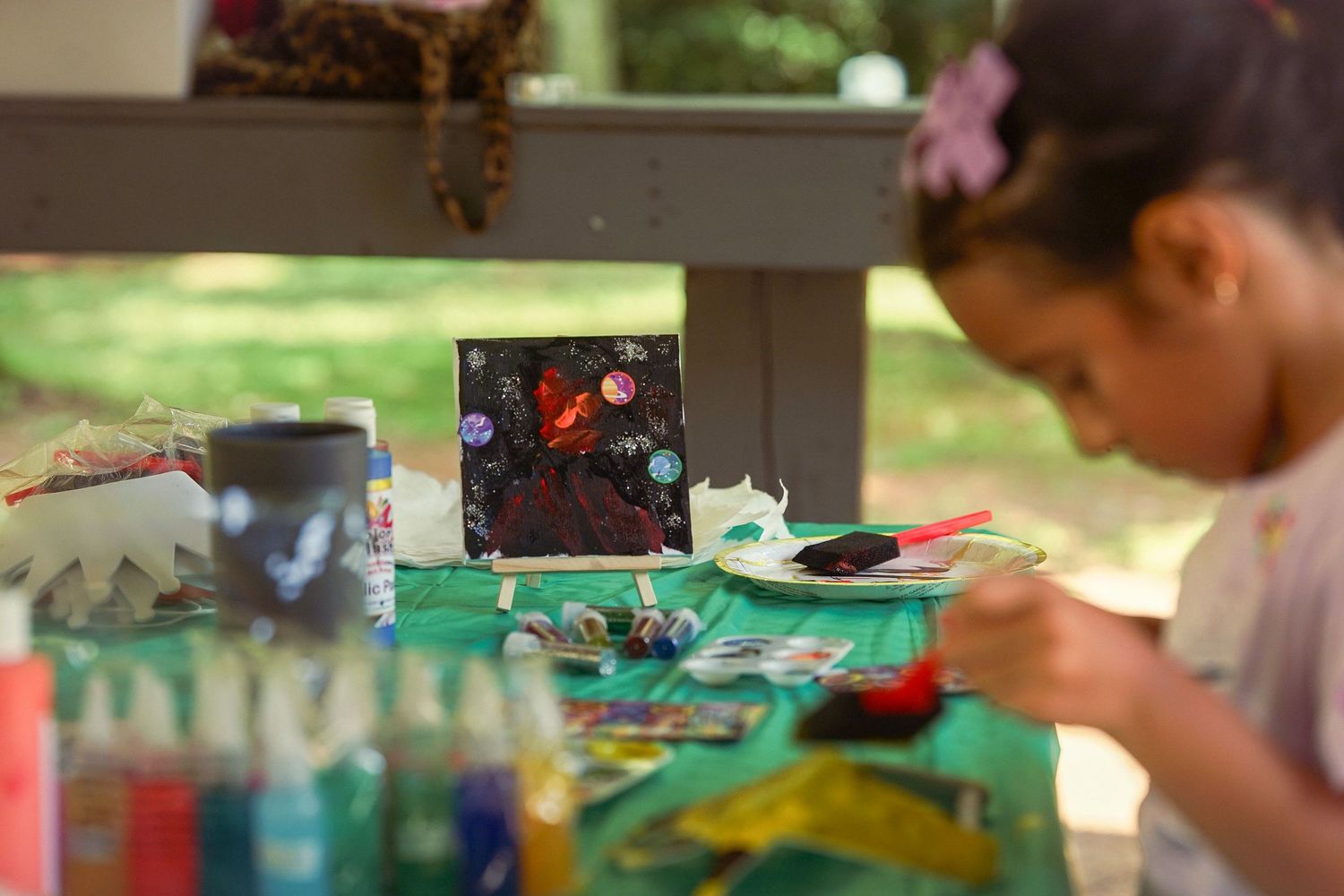 Young girl painting a colorful canvas at an outdoor art table with supplies scattered around.