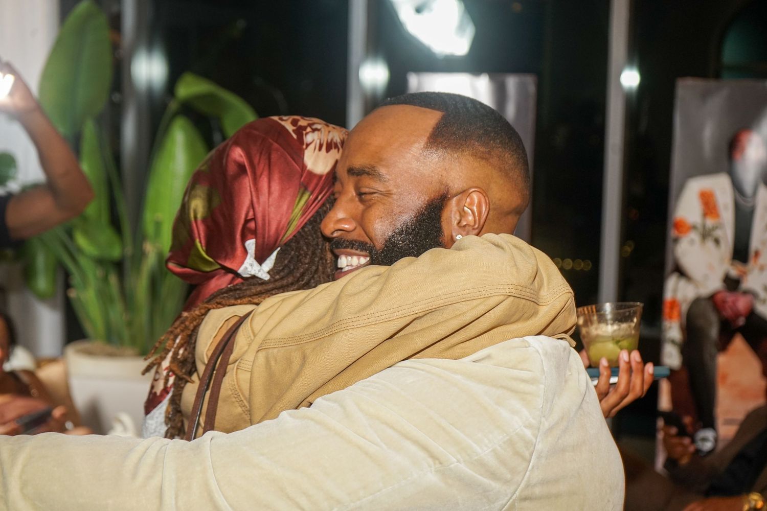 Two people sharing a warm embrace at an indoor gathering, smiling joyfully in a cozy, plant-filled setting.