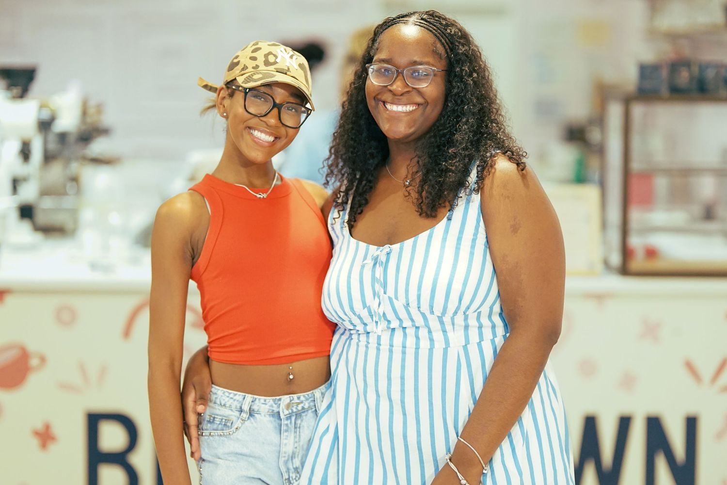 Two smiling women posing together inside a bright cafe, one wearing an orange crop top and the other a blue striped dress.