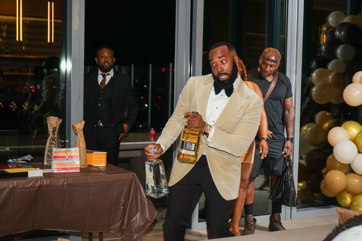 Man in gold blazer and bow tie holds whiskey bottle at elegant black and gold gala event with guests.