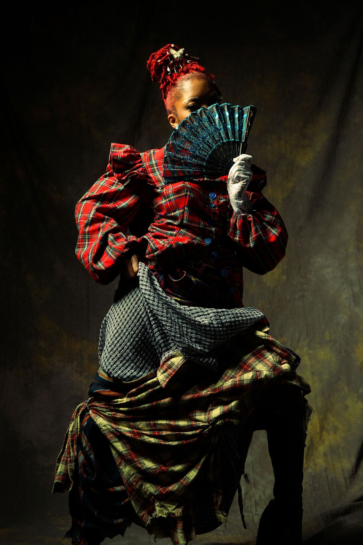 A dramatic portrait in dark lighting shows a figure in mixed plaid patterns holding a decorative blue fan against a moody background.