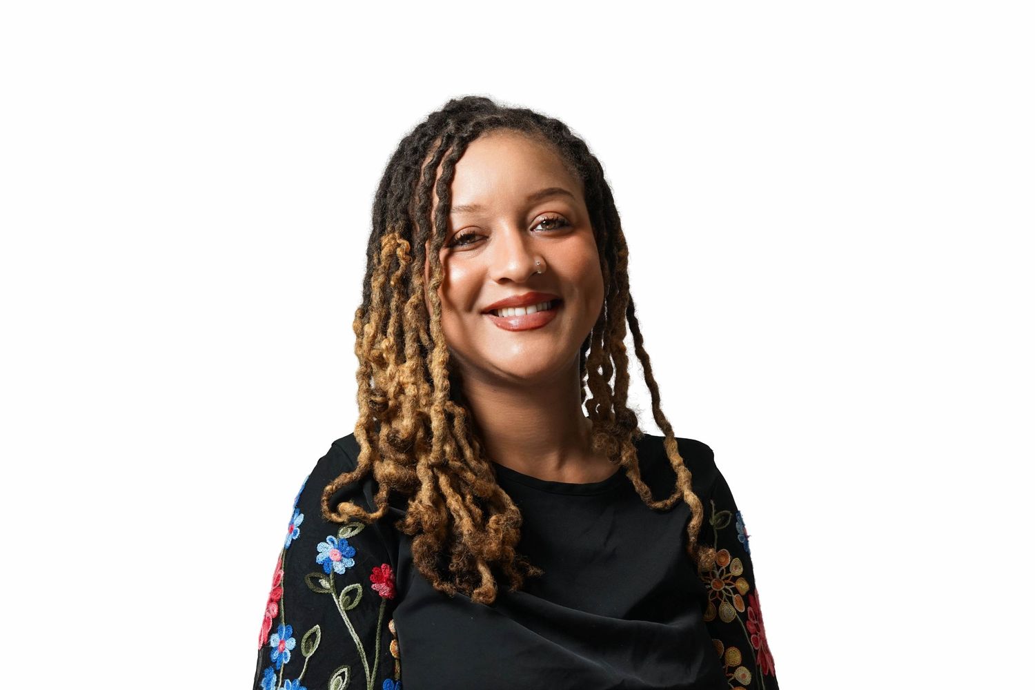 Smiling woman with long locs wearing a black top and floral embroidered jacket against white background.