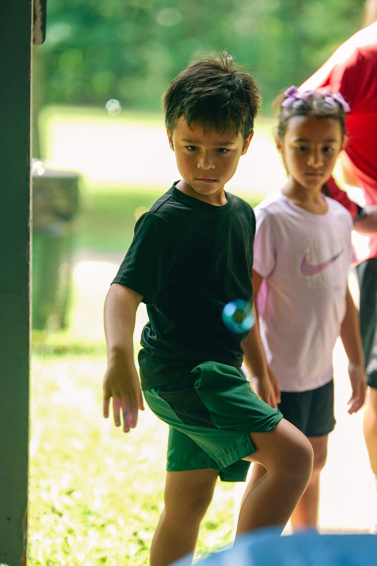 Young boy in black shirt and green shorts playing outdoors with a girl in pink Nike shirt behind him.