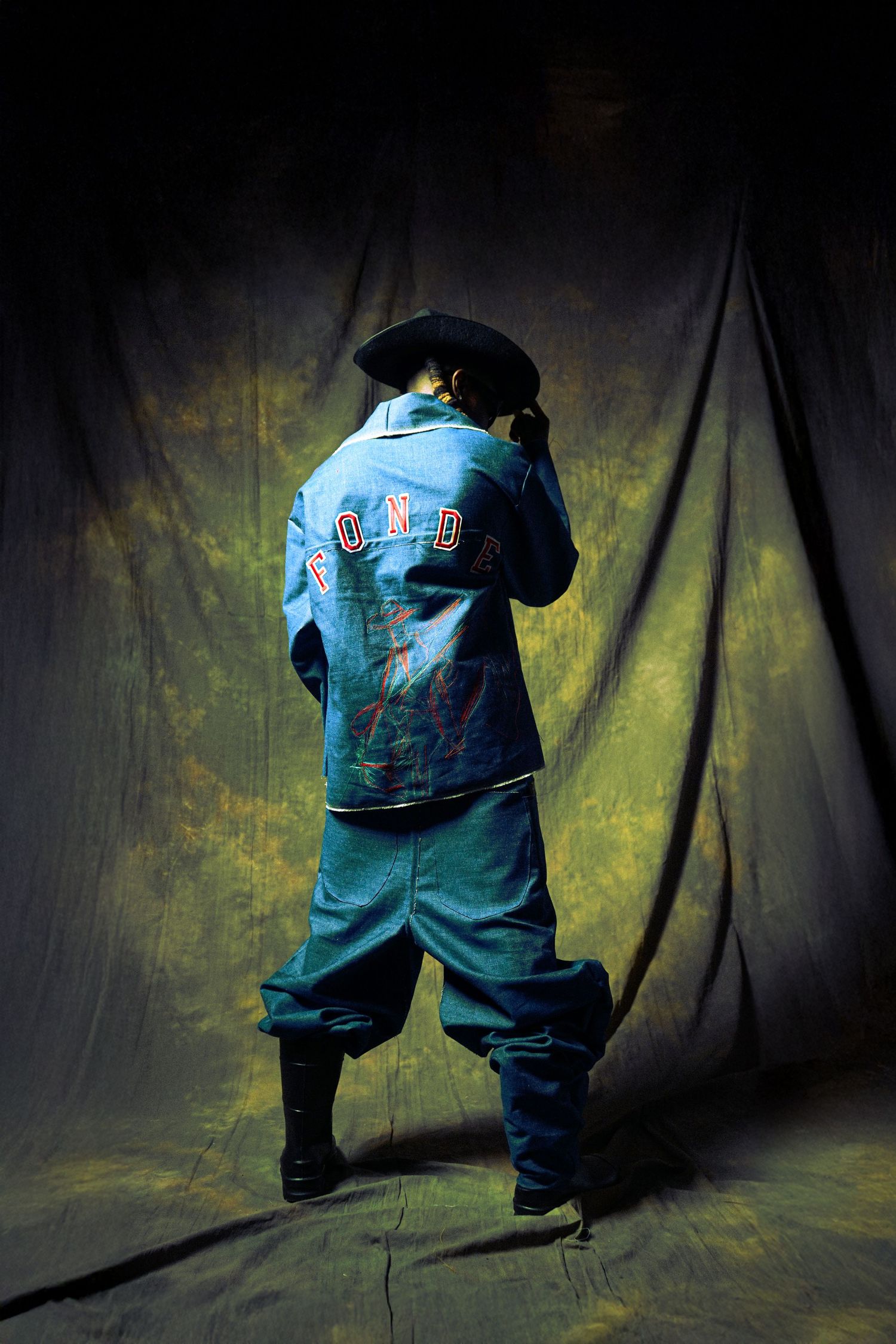 Figure in baggy turquoise outfit and hat poses against dark backdrop with dramatic lighting and yellow tones.