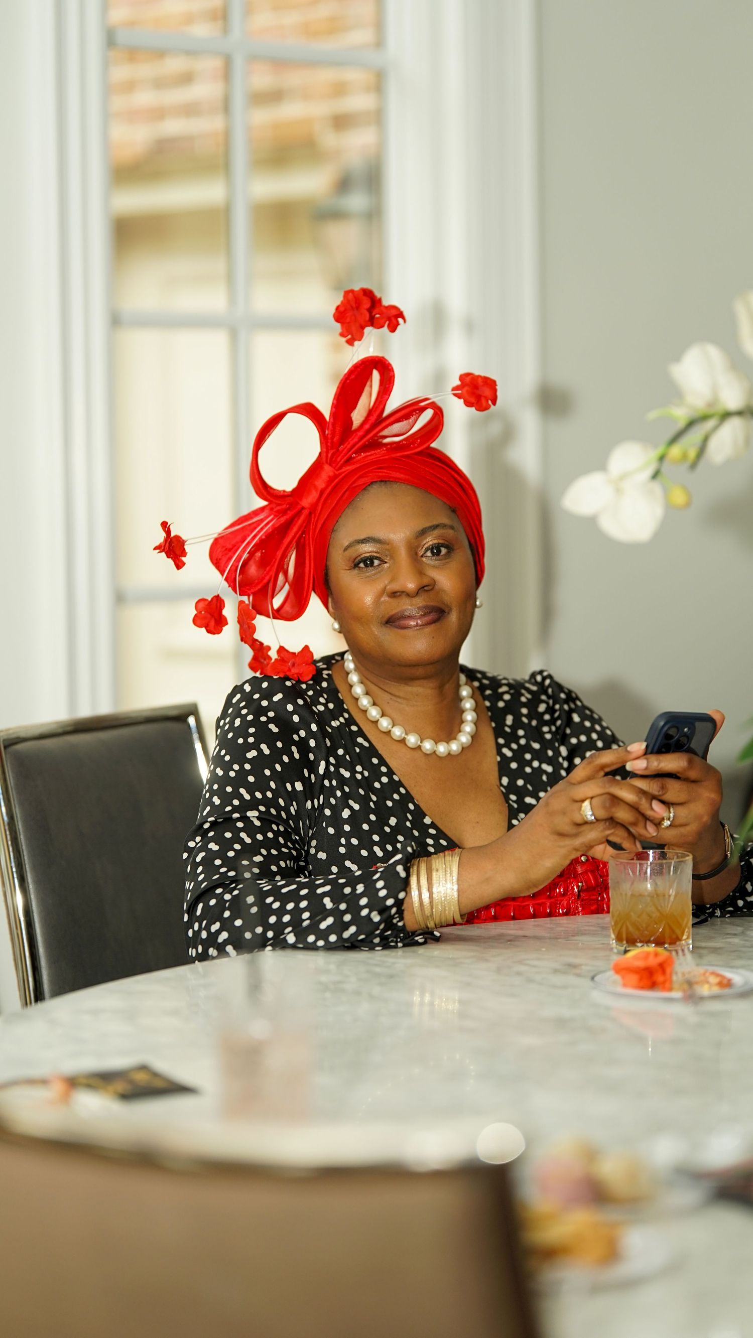 Person in polka dot dress and red fascinator sitting at dining table with phone and beverage.
