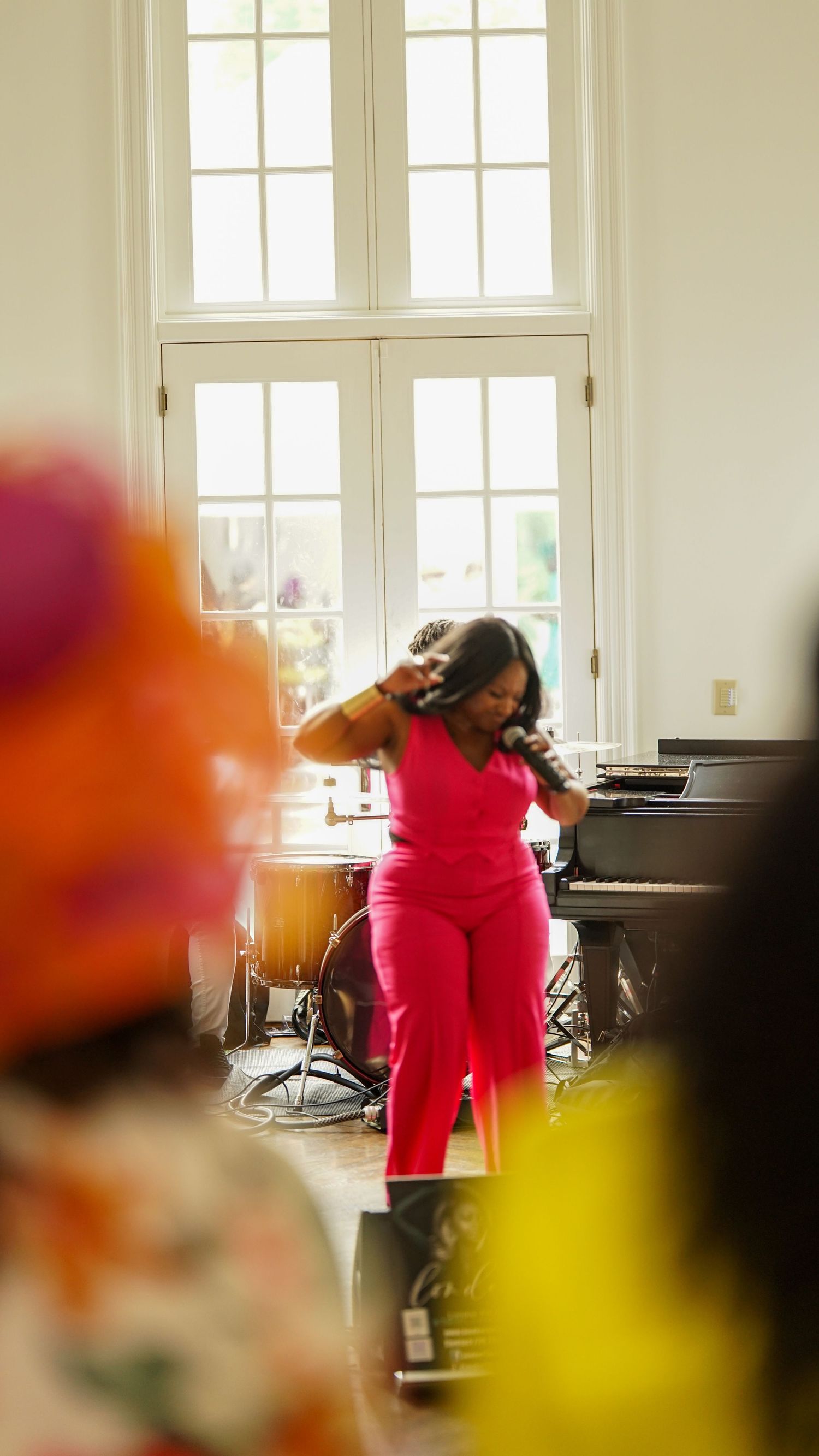 Performer in bright red outfit singing on stage with piano and band setup in bright windowed room.