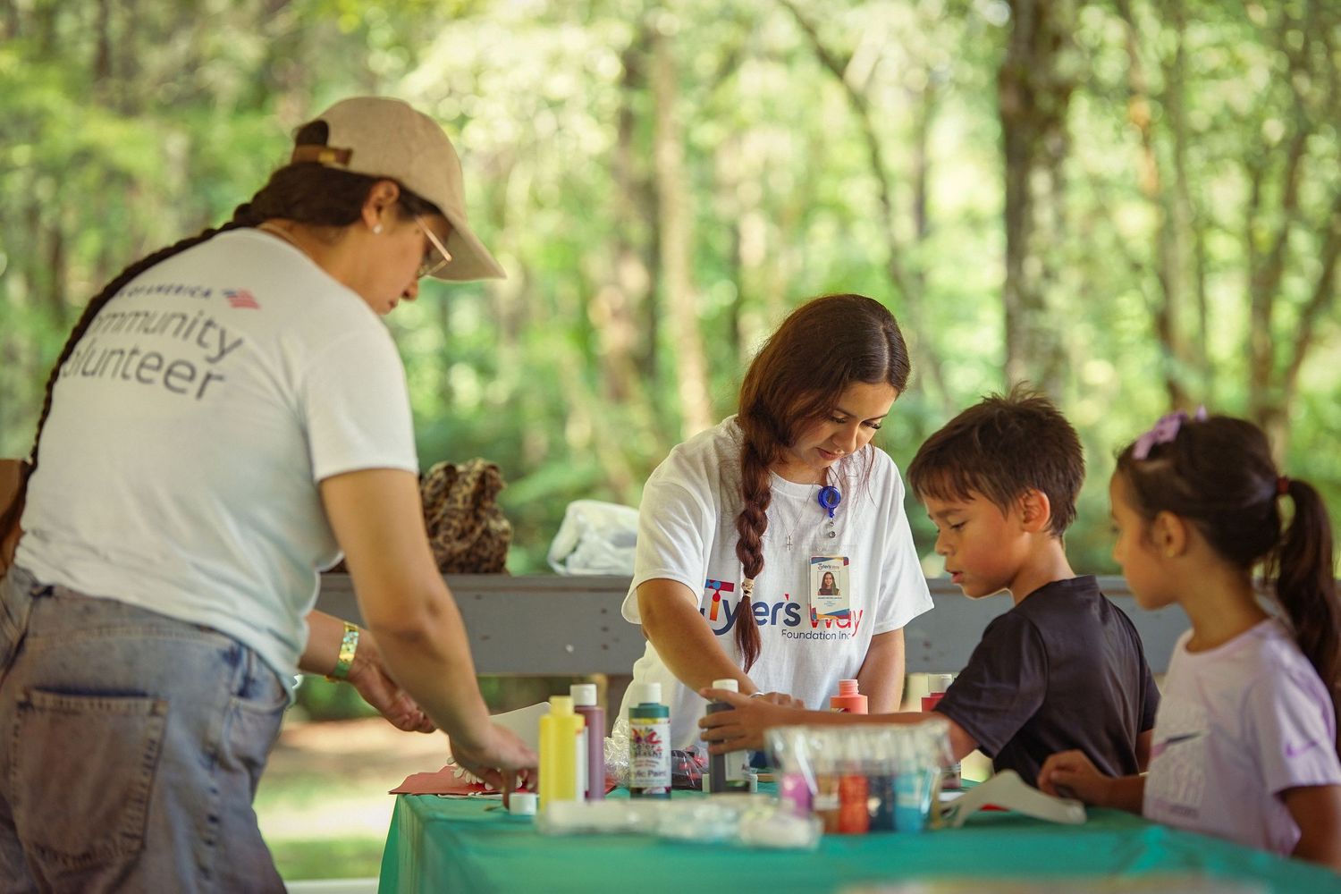 Community volunteers engage with children at an outdoor craft table surrounded by lush green trees.
