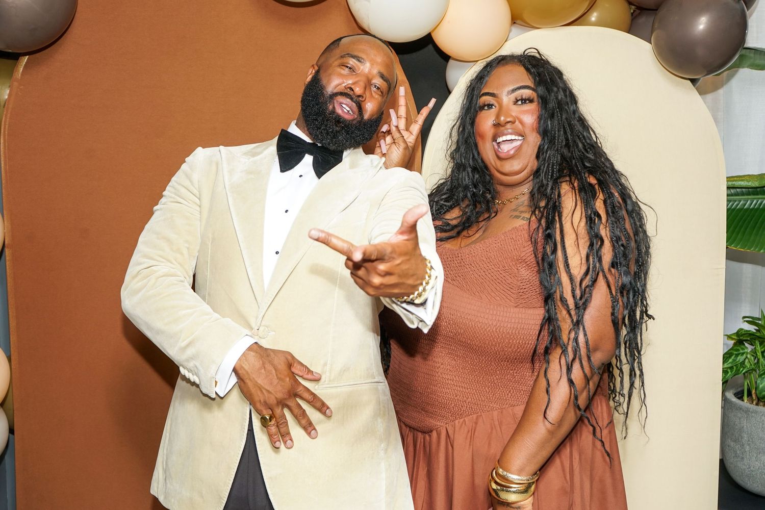 A man in a white tuxedo and a woman in a rust dress pose playfully at a festive event with balloon decor.