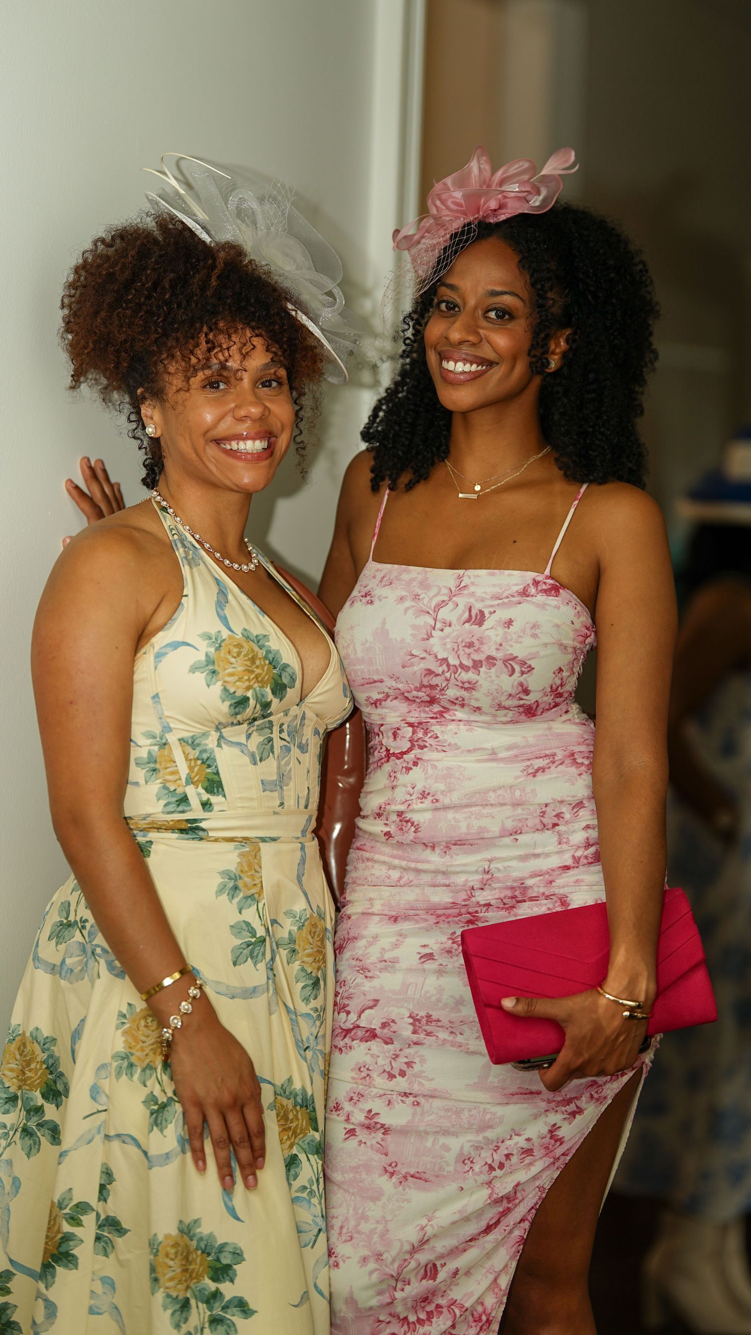 Two people in floral dresses and feathered fascinators pose together at an indoor party.