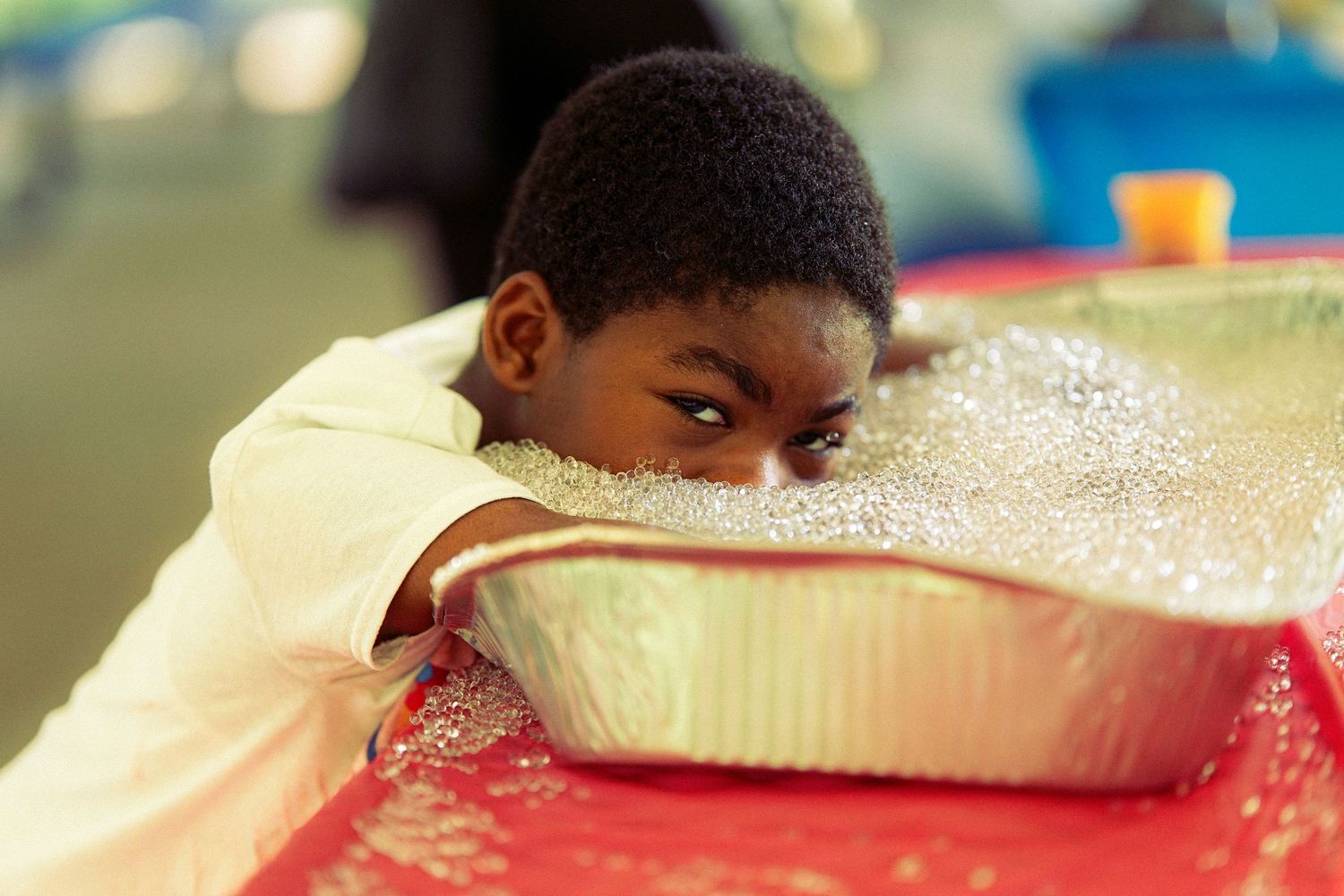 A young boy rests his chin on the edge of a foil pan at an outdoor activity table.