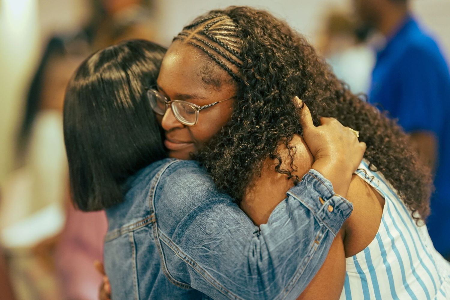 Two women share a warm emotional hug at an indoor community gathering event.