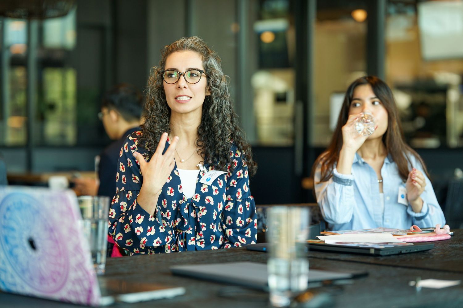 A curly-haired woman with glasses speaks expressively during a business meeting at a cafe.