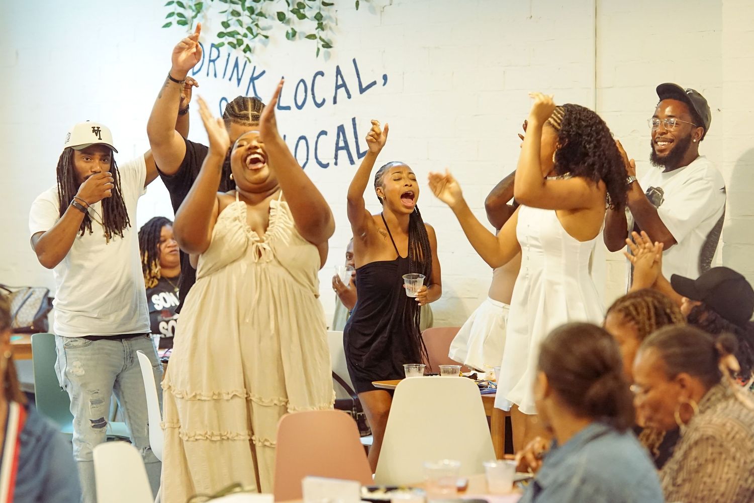 Joyful group of Black people celebrating and cheering inside a venue with a Drink Local sign in background.