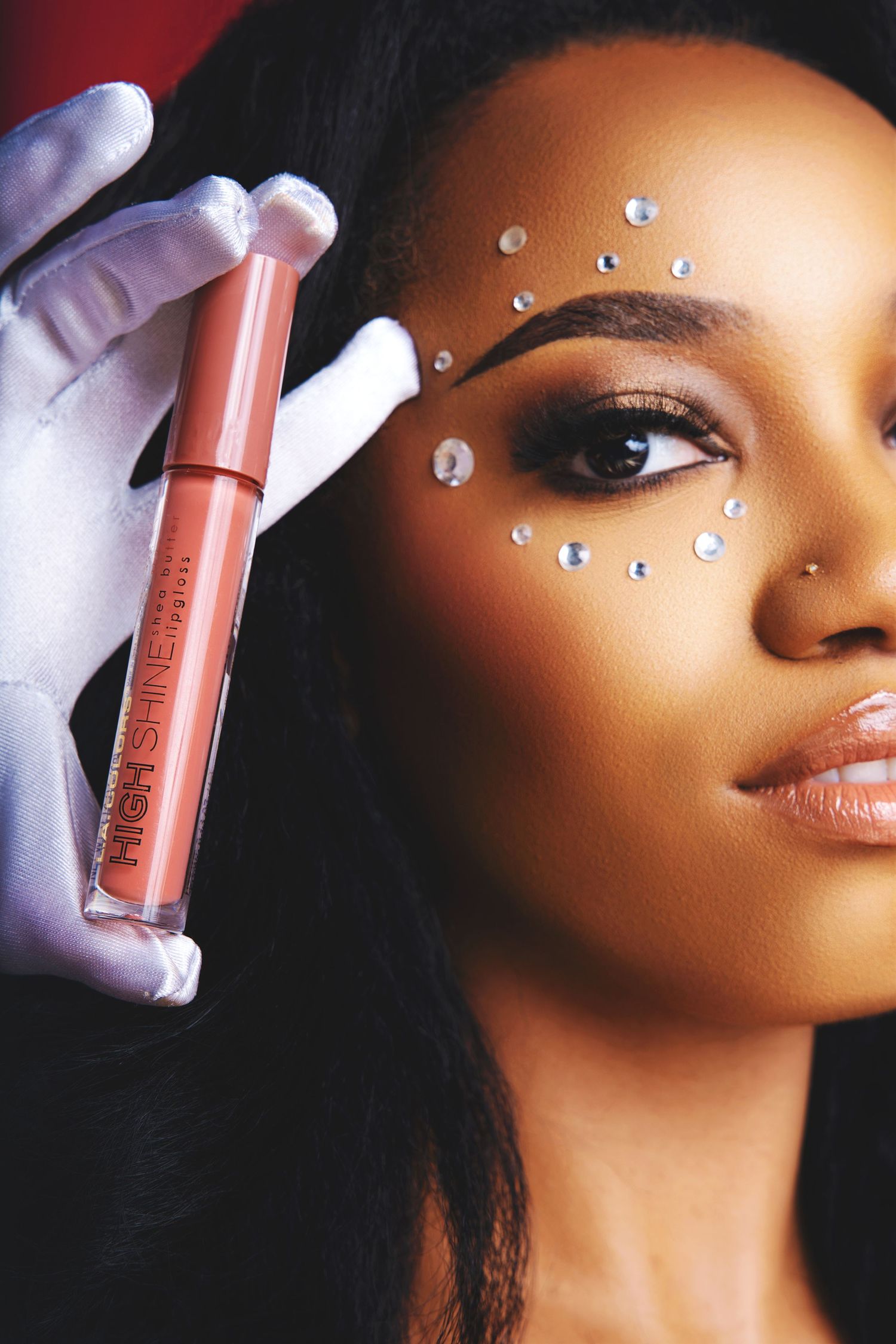 Beauty shot featuring crystal face gems and coral liquid lipstick being held by gloved hand.