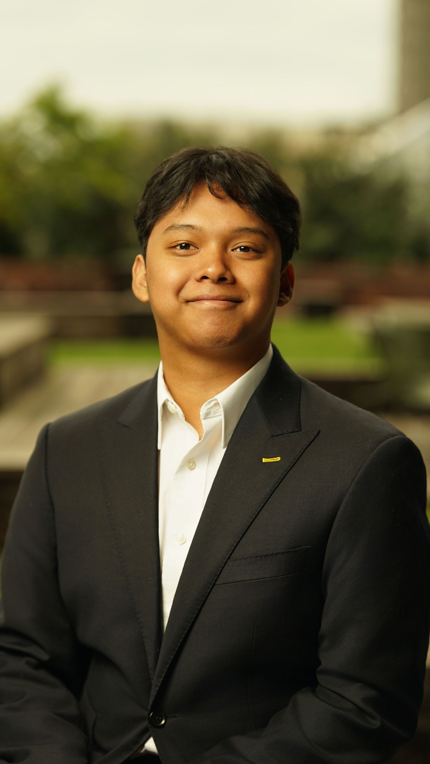 Professional in dark suit and white shirt poses outdoors with friendly smile.