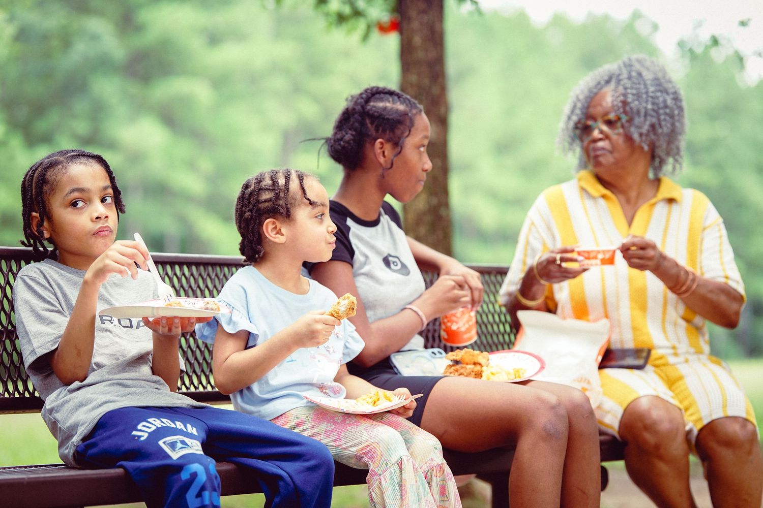 Family enjoying a meal together on a park bench outdoors, with children and grandmother sharing food.