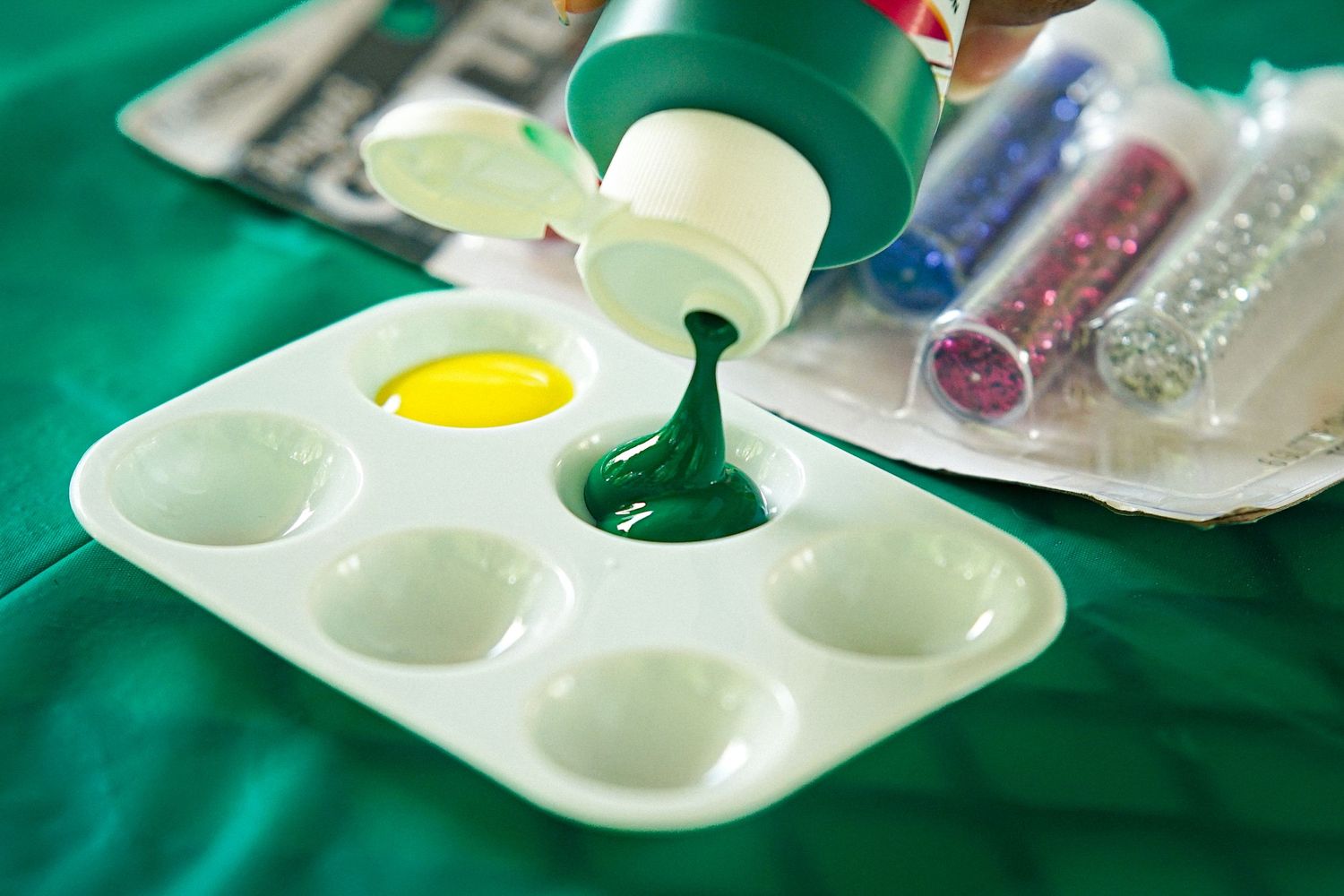 Green paint being squeezed onto a white palette tray with yellow paint, glitter tubes nearby on green fabric.