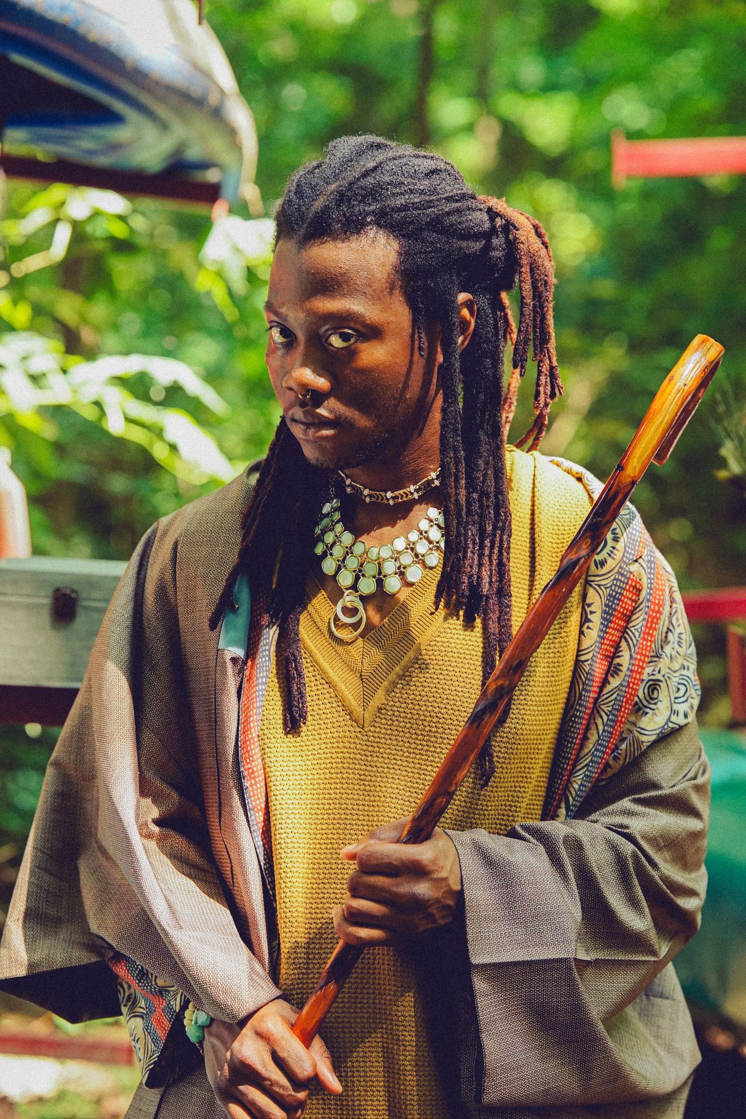 A man with dreadlocks wearing colorful layered clothing and a beaded necklace holds a wooden staff outdoors.