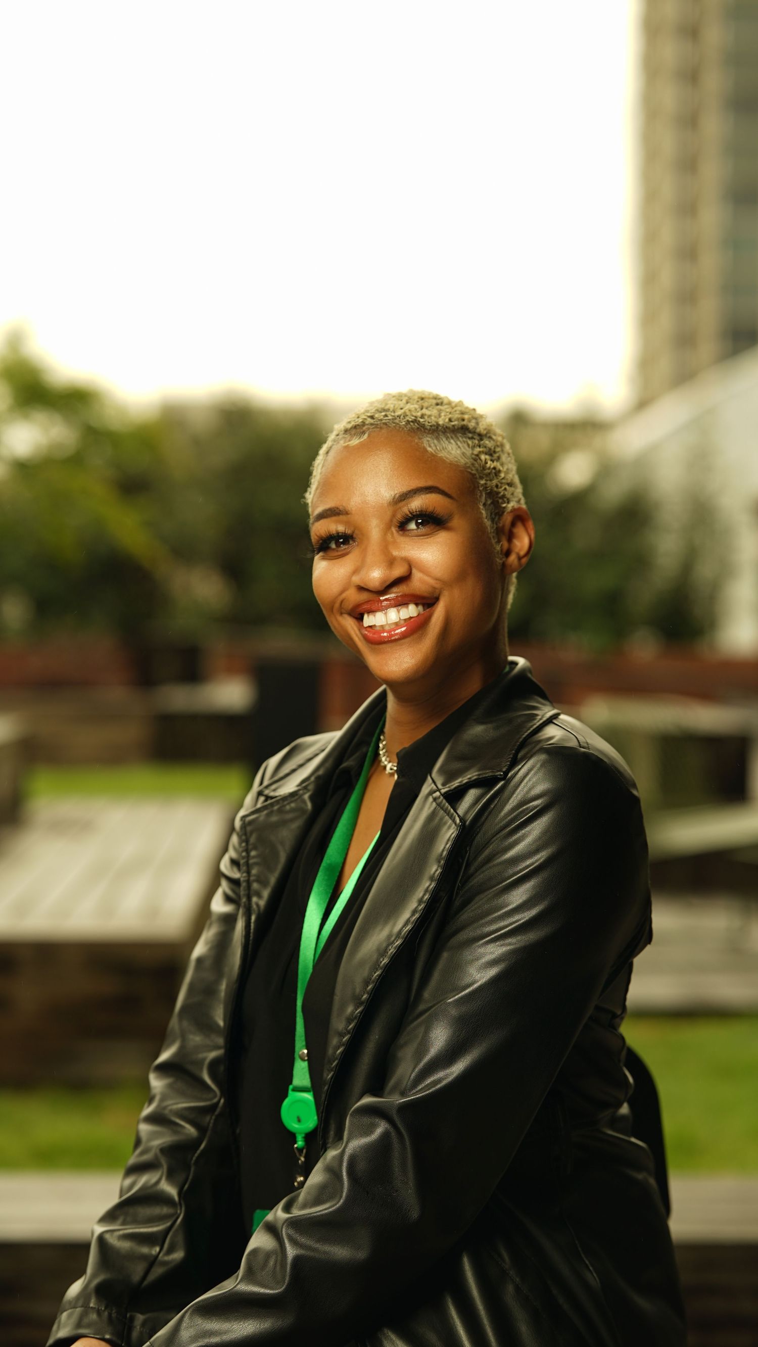 Person in leather jacket and green necklace smiling warmly during an outdoor photoshoot.